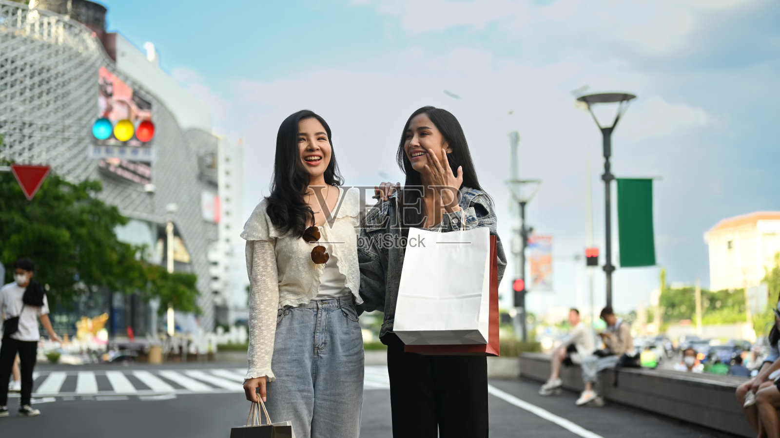 时尚的女性朋友们拿着购物袋站在城市大道上,背景是模糊的城市景观和蓝天。照片摄影图片