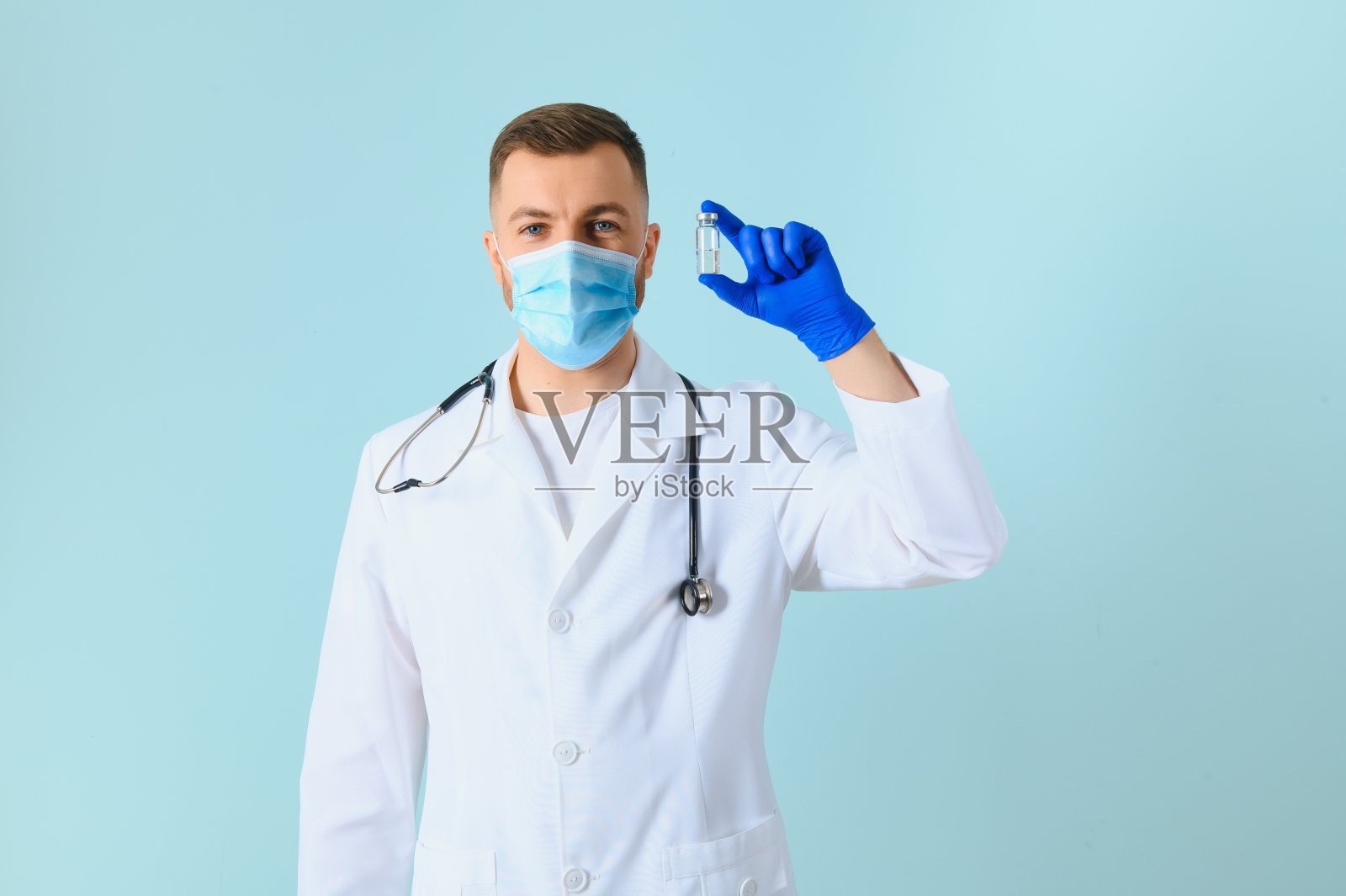 年轻的医生戴着医用口罩和白色外套,听诊器拿着注射器隔离在蓝色上照片摄影图片