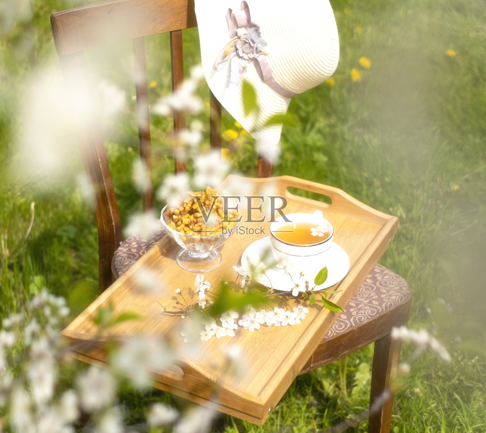 在一个阳光明媚的日子,在樱桃园吃着茶点,在乡村度过暑假。在鲜花和果树之间,有一个托盘和一顶帽子的椅子照片摄影图片