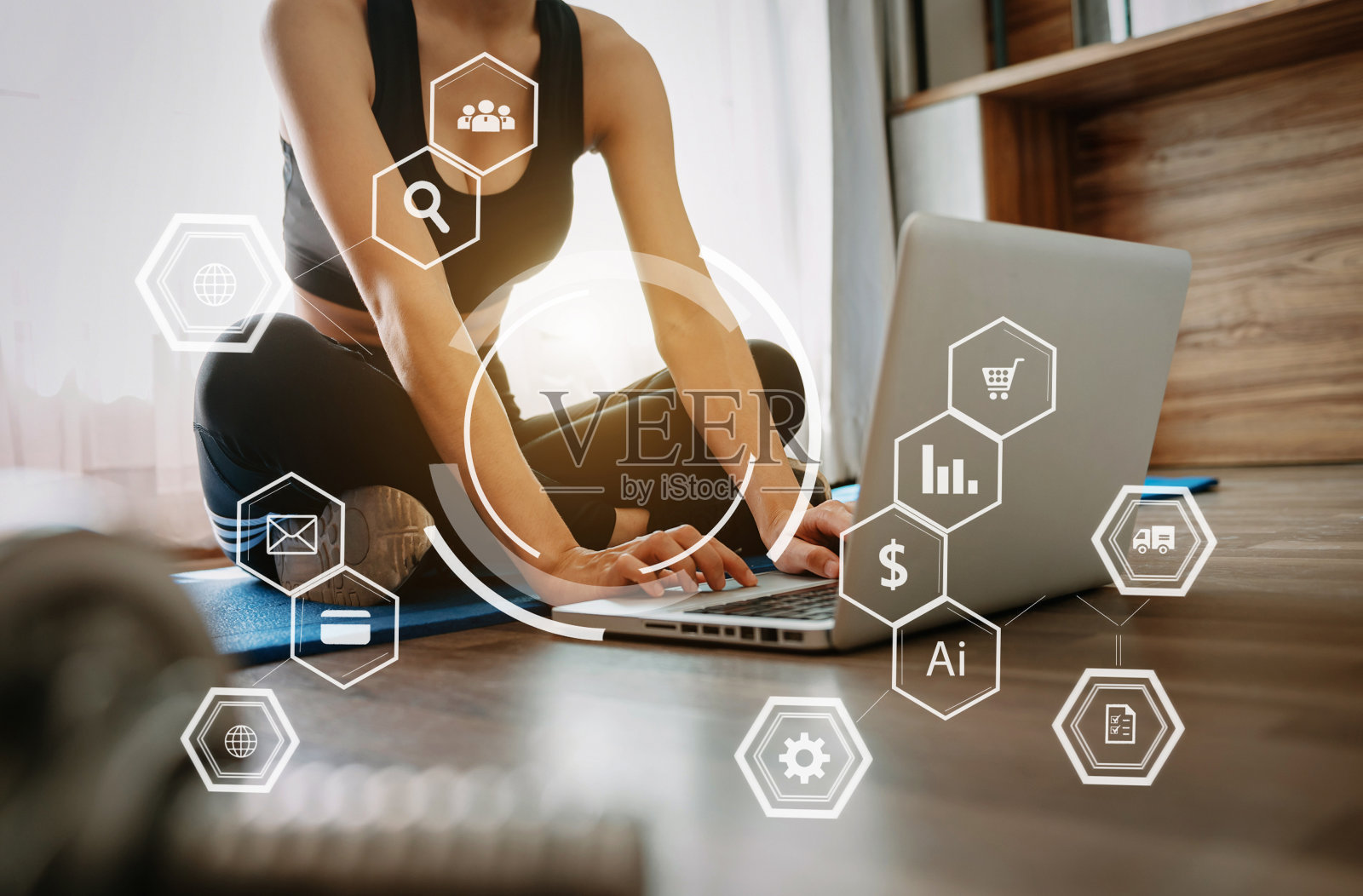 一位穿着运动服的运动女性坐在地板上,手里拿着哑铃,喝着蛋白奶昔或一瓶水,正在使用笔记本电脑。运动和娱乐概念。照片摄影图片