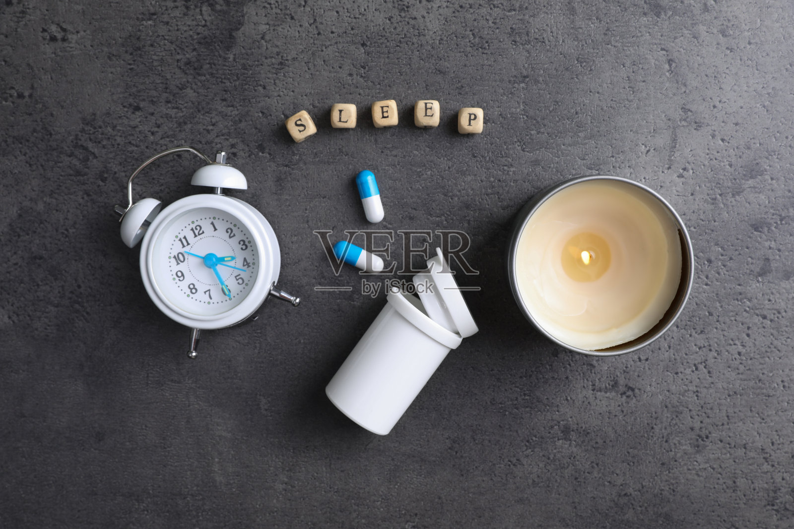 单词Sleep由木制的立方体制成,靠近闹钟,蜡烛和药丸在灰色的背景上,平躺着。失眠的治疗照片摄影图片