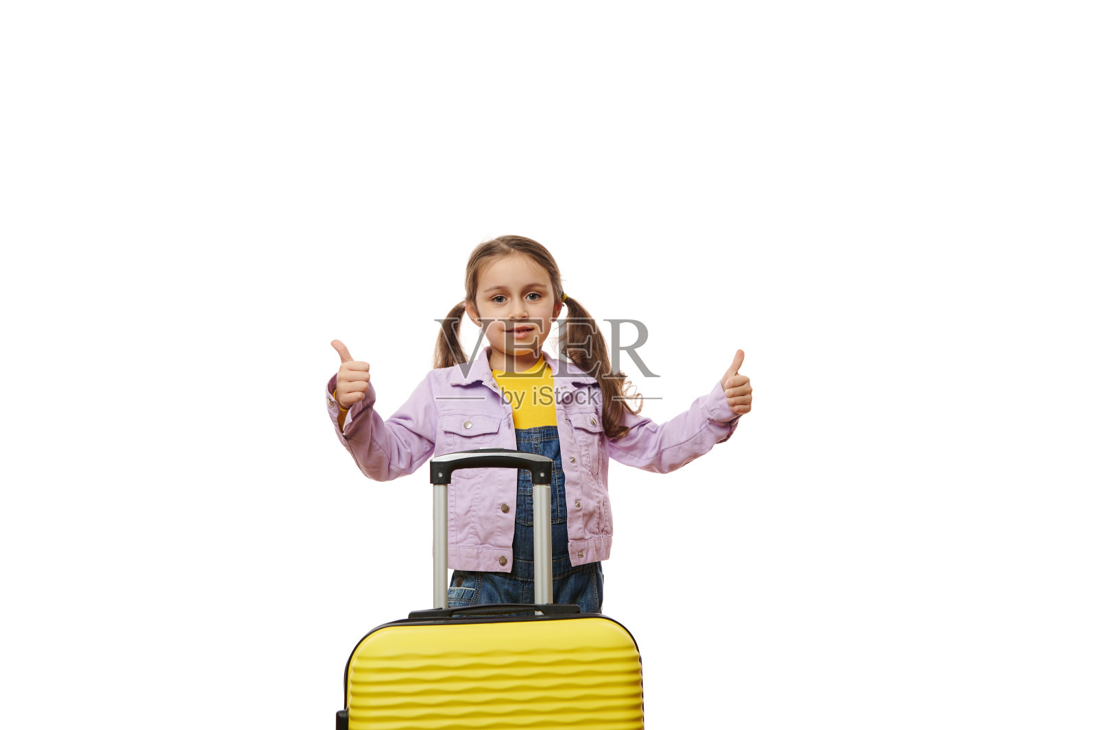 可爱的旅行者女婴扎着两个马尾辫,站在一个黄色的行李箱后面,竖起大拇指看着镜头照片摄影图片