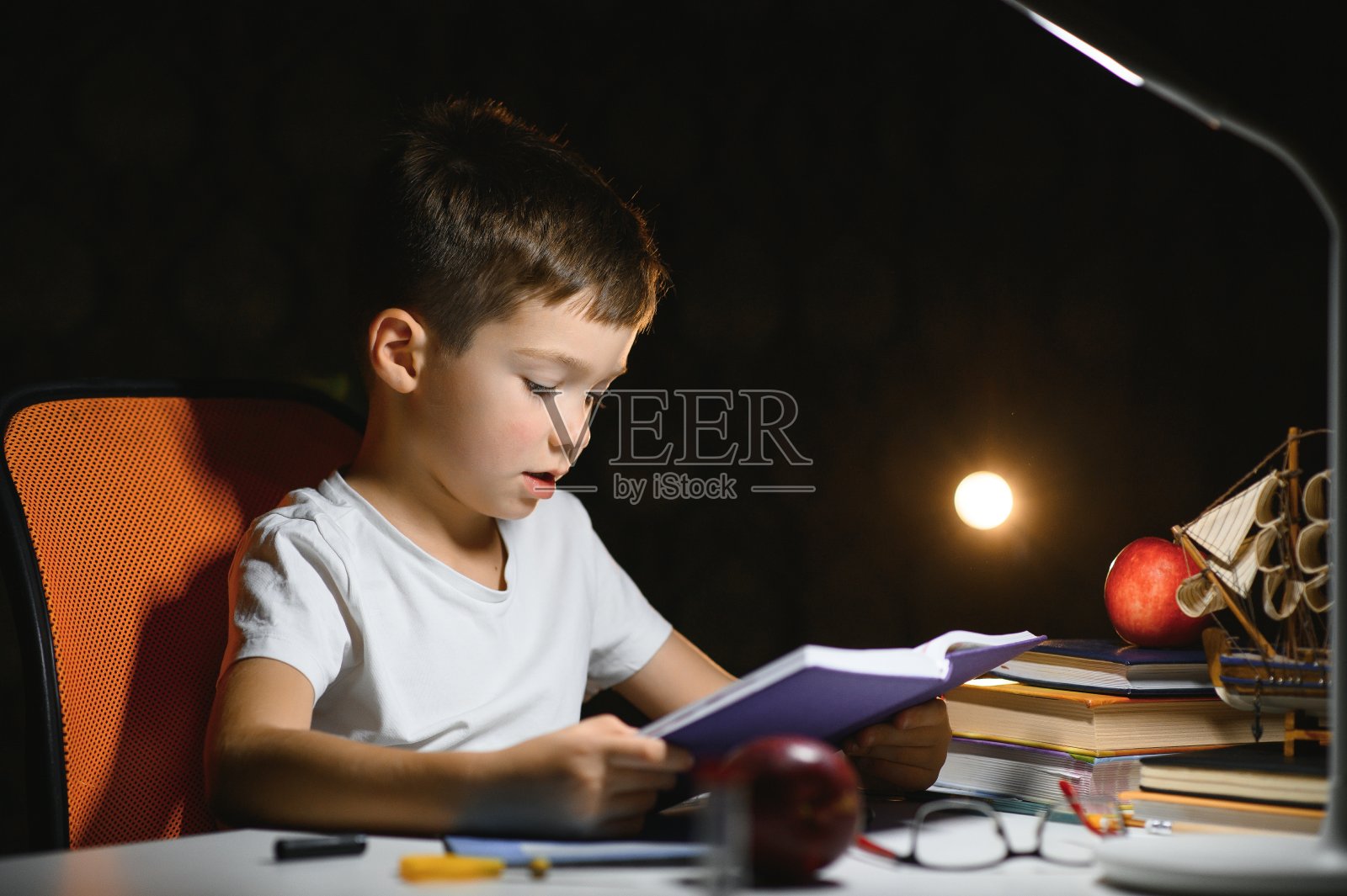 男孩在家里学习功课,在台灯的灯光下坐在桌子旁。照片摄影图片