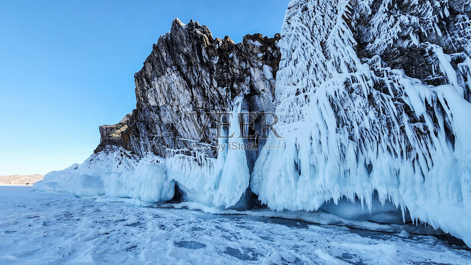 冬季山地景观。湖上的岩石上结满了冰和冰柱。湖上的海角是一个晴朗的冬日,有蓝天,有云,有太阳。冬天贝加尔湖上的岩石。照片摄影图片