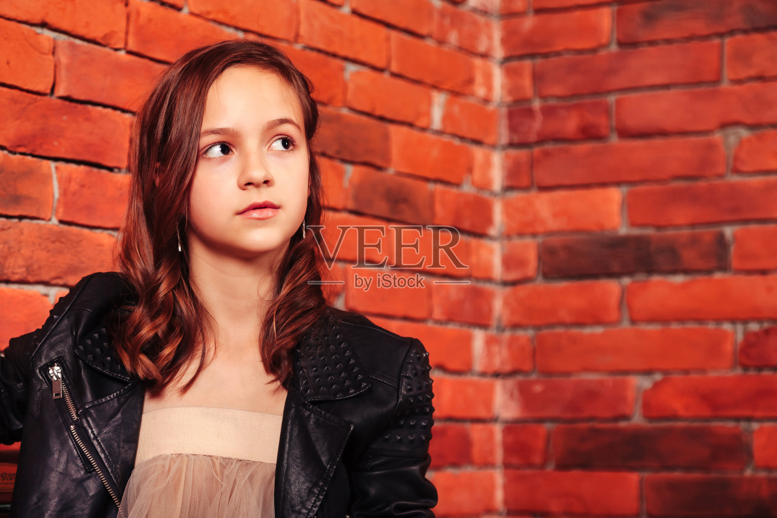 肖像少年女孩在皮夹克坐在长凳在砖墙背景,看向别处。可爱的少女模特等待和摆造型的街道室外。时尚风格概念。复制文本空格照片摄影图片