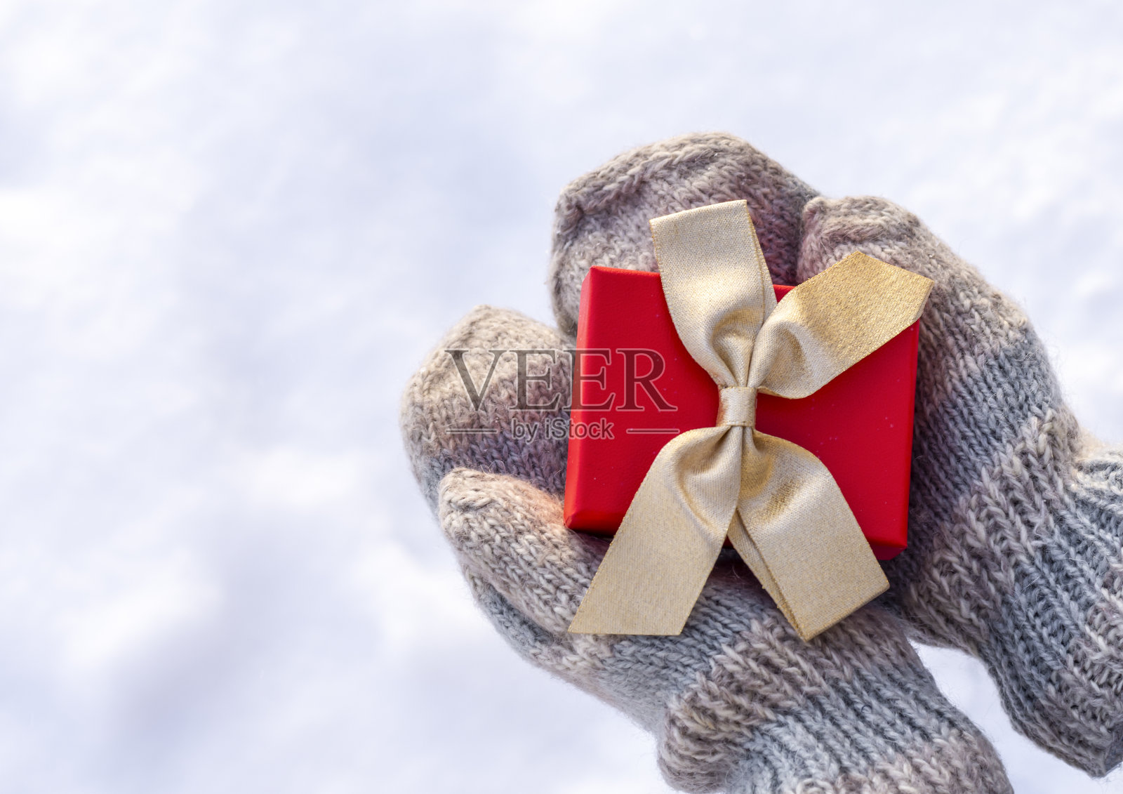 女人戴着浅灰色针织手套的手拿着一个漂亮的礼物,盒子里有一个金色的蝴蝶结,背景是一个下雪的冬天。爱情的概念,情人,恭喜你。照片摄影图片