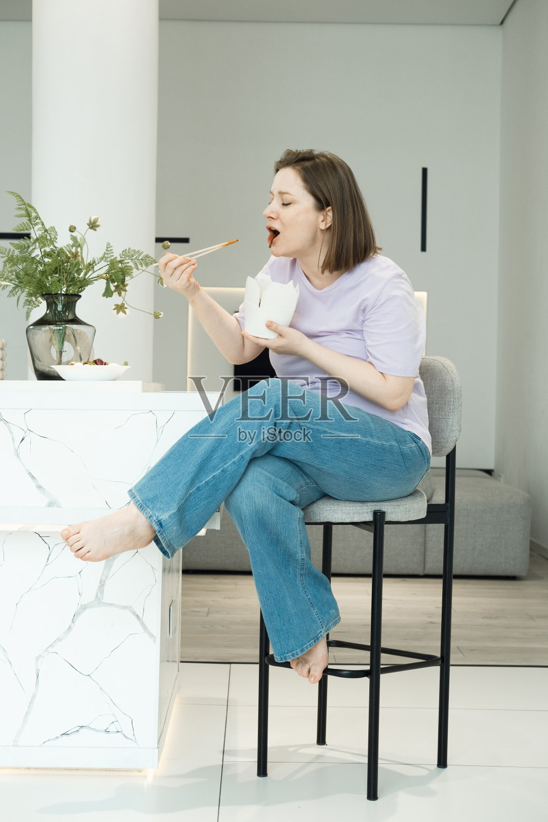 年轻的胖女人坐在厨房里,吃着白色纸盒里的面条。食品交付。不健康的食物。照片摄影图片