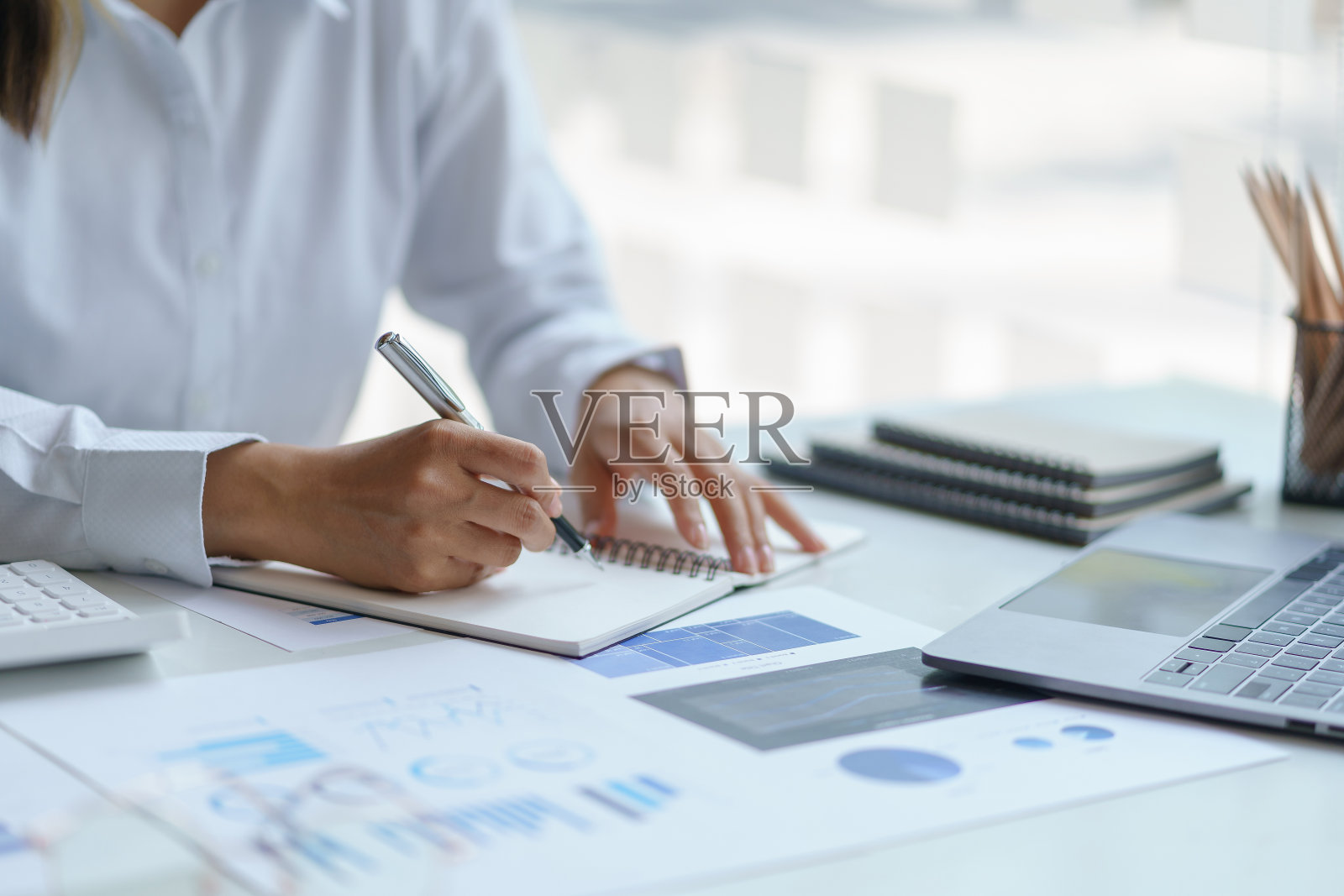 一个女人在办公室用笔记本电脑在笔记本上写字和记笔记的特写图像。照片摄影图片