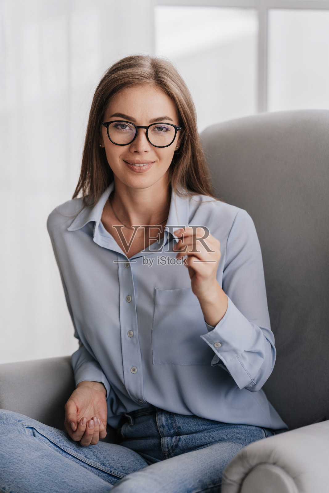 垂直拍摄自信的意大利金发年轻女子坐在椅子上,戴着眼镜,穿着蓝色衬衫和蓝色牛仔裤,微笑着看着家里的相机。迷人的瑞典家庭主妇在周末放松。照片摄影图片