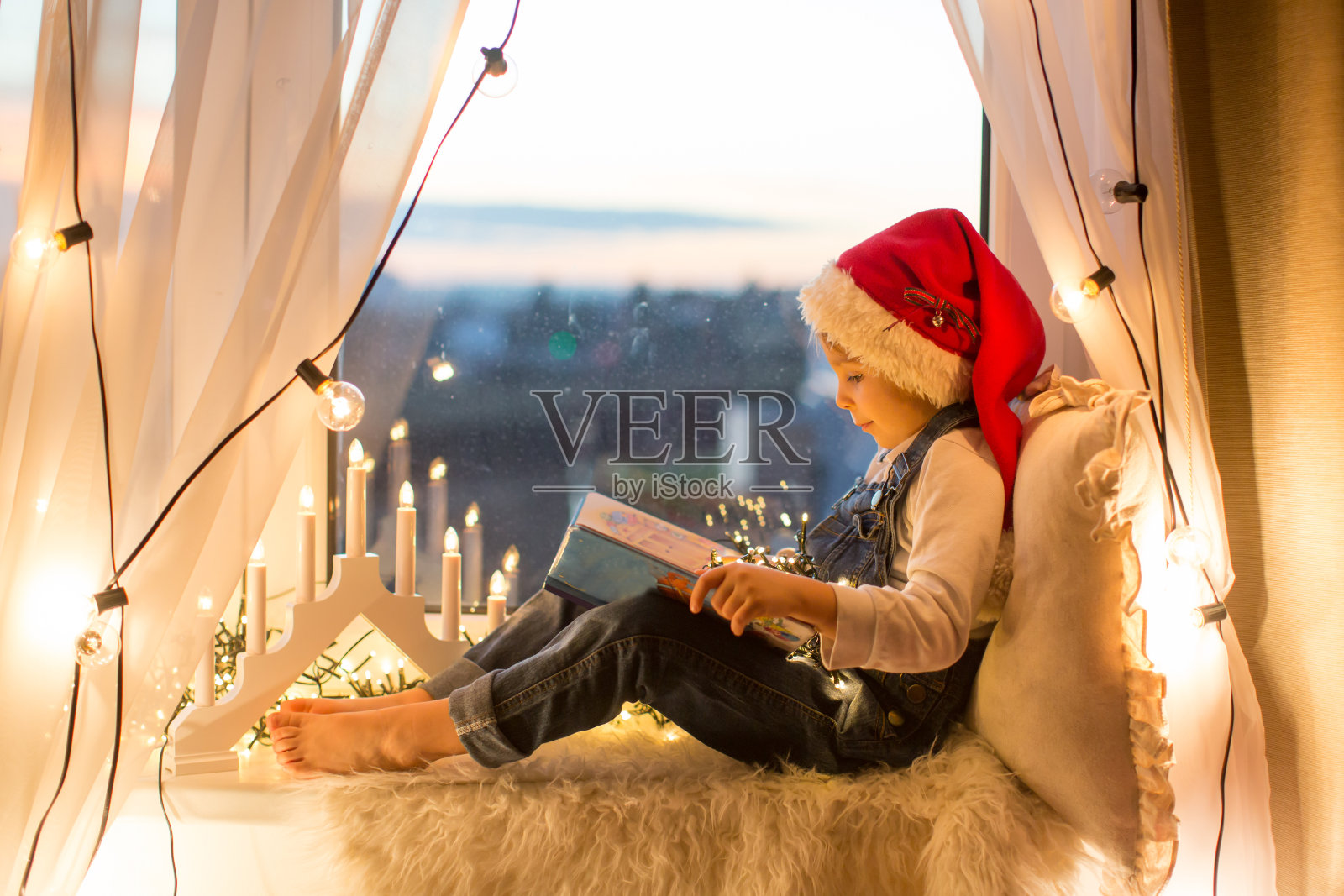 蹒跚学步的孩子,可爱的金发男孩,坐在窗户上,看书,周围是圣诞彩灯照片摄影图片