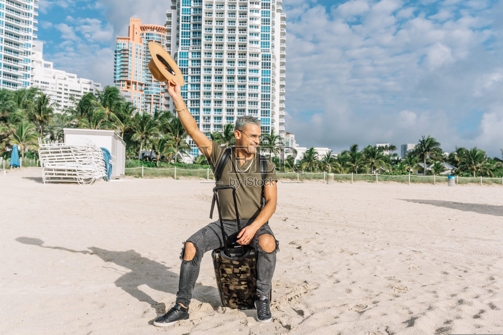 戴眼镜的男人,坐在手提行李上的男人,快乐地挥舞着他的帽子照片摄影图片