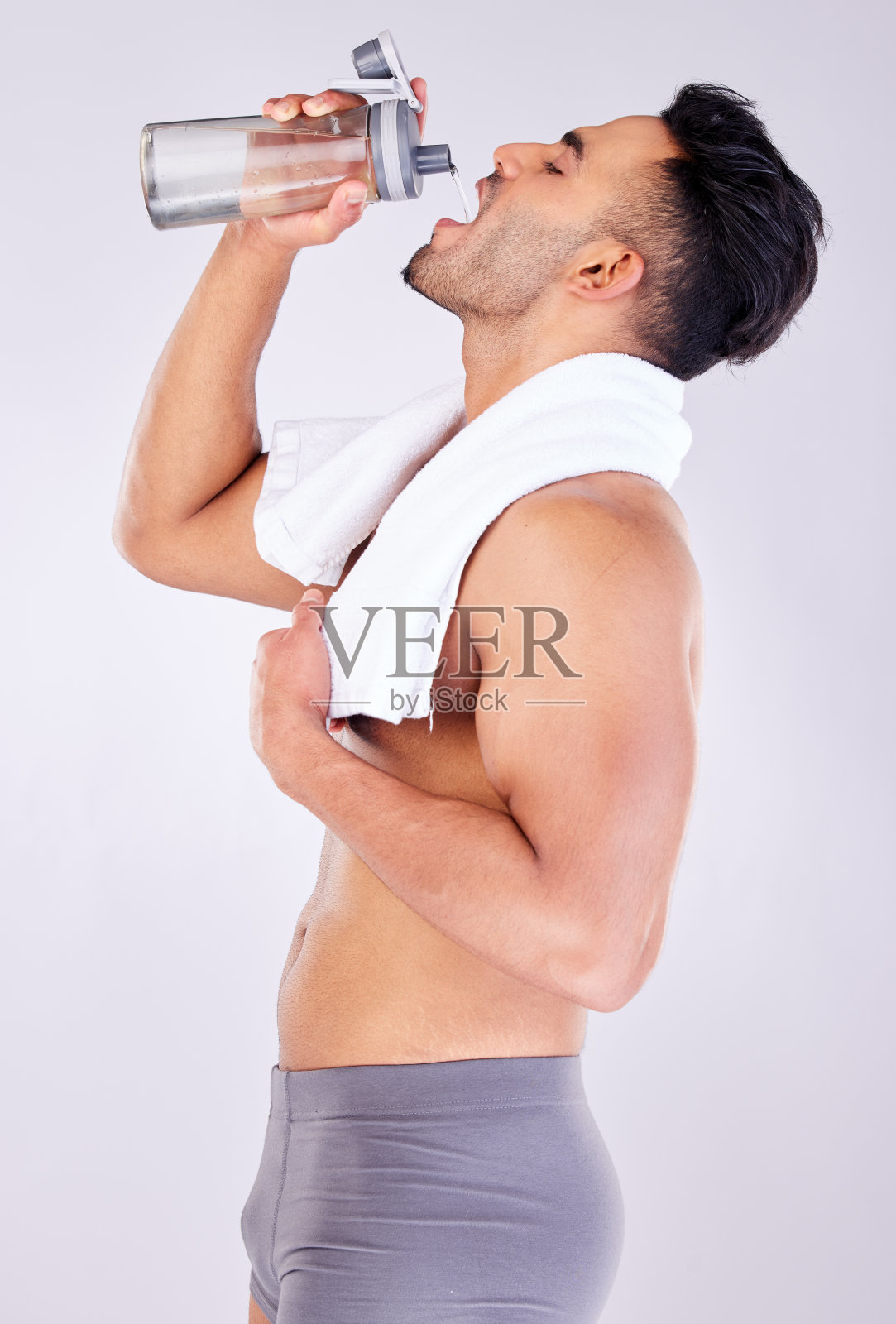 男士在健身房锻炼或淋浴后健身训练后用瓶子、内衣和毛巾喝水。健康,健康和水合作用,私人教练站着喝水与工作室背景照片摄影图片