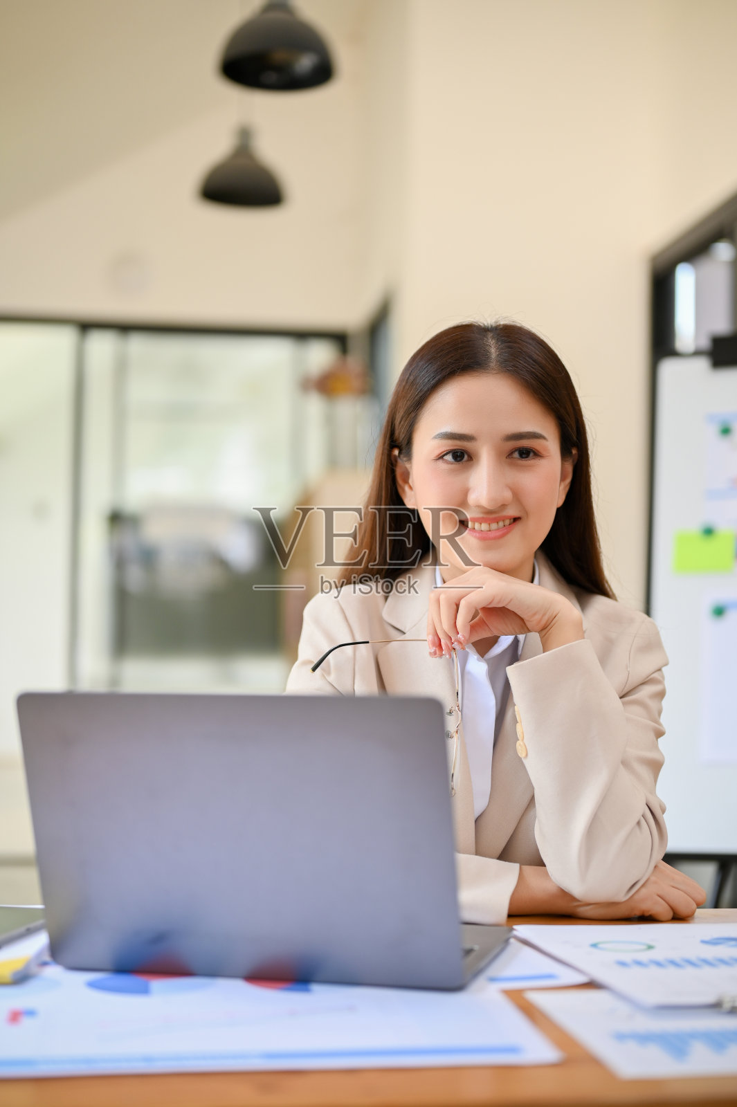 迷人的亚洲女商人坐在她的办公桌前,手放在下巴上,微笑着看着镜头。照片摄影图片