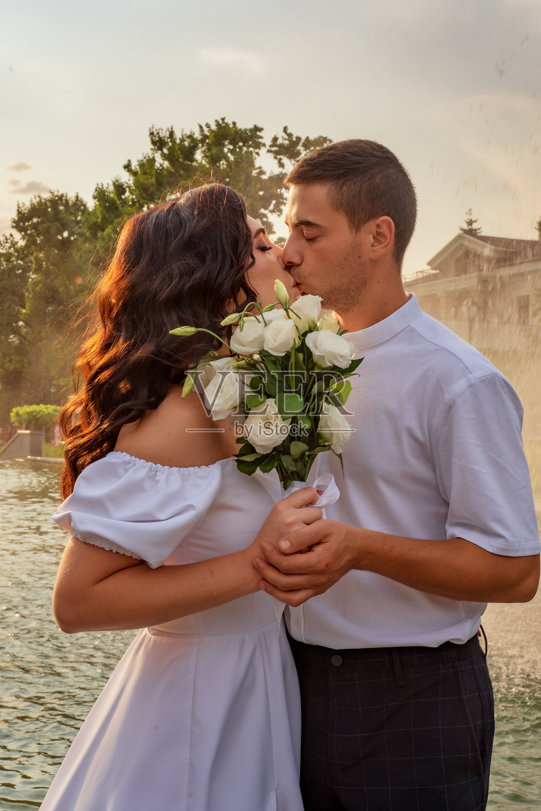 年轻人在纽约中央公园热烈接吻。年轻情侣接吻特写。接吻情侣的肖像。男孩和女孩穿着白色的衣服,手捧一束白色的玫瑰,温柔地接吻。照片摄影图片