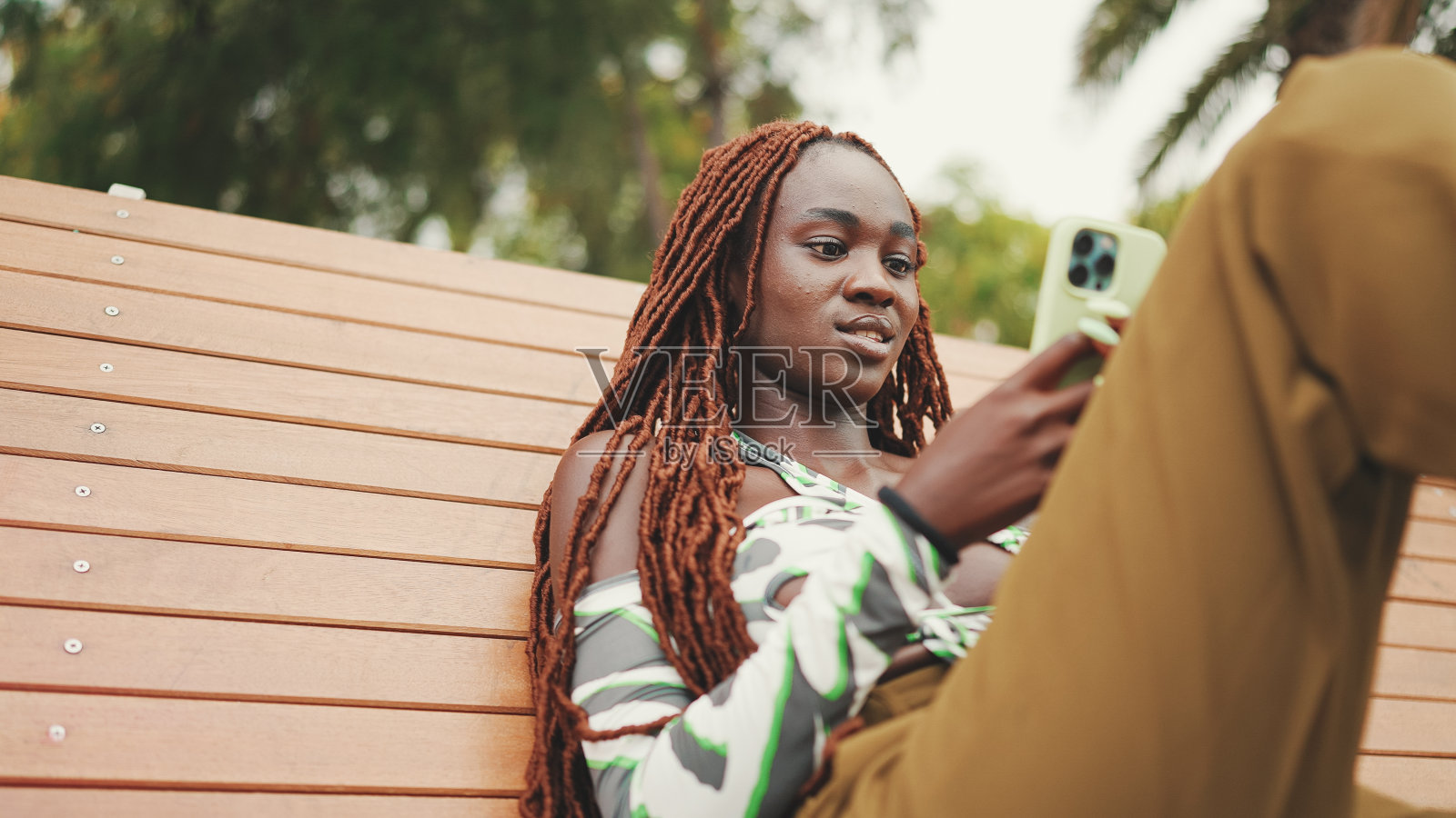 微笑的华丽女子与非洲辫子穿着在顶部是使用手机,而坐在长凳上。时髦的女孩看照片,视频在她的智能手机照片摄影图片