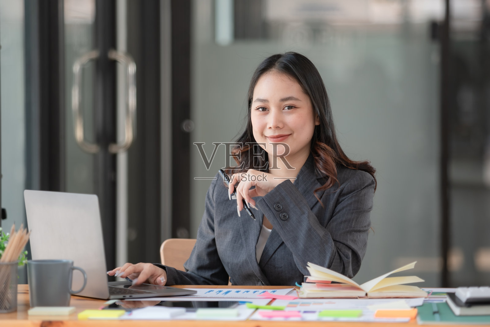 一个年轻的女商人使用笔记本电脑的肖像,微笑着看着相机,坐在一个现代办公室的办公桌前。照片摄影图片
