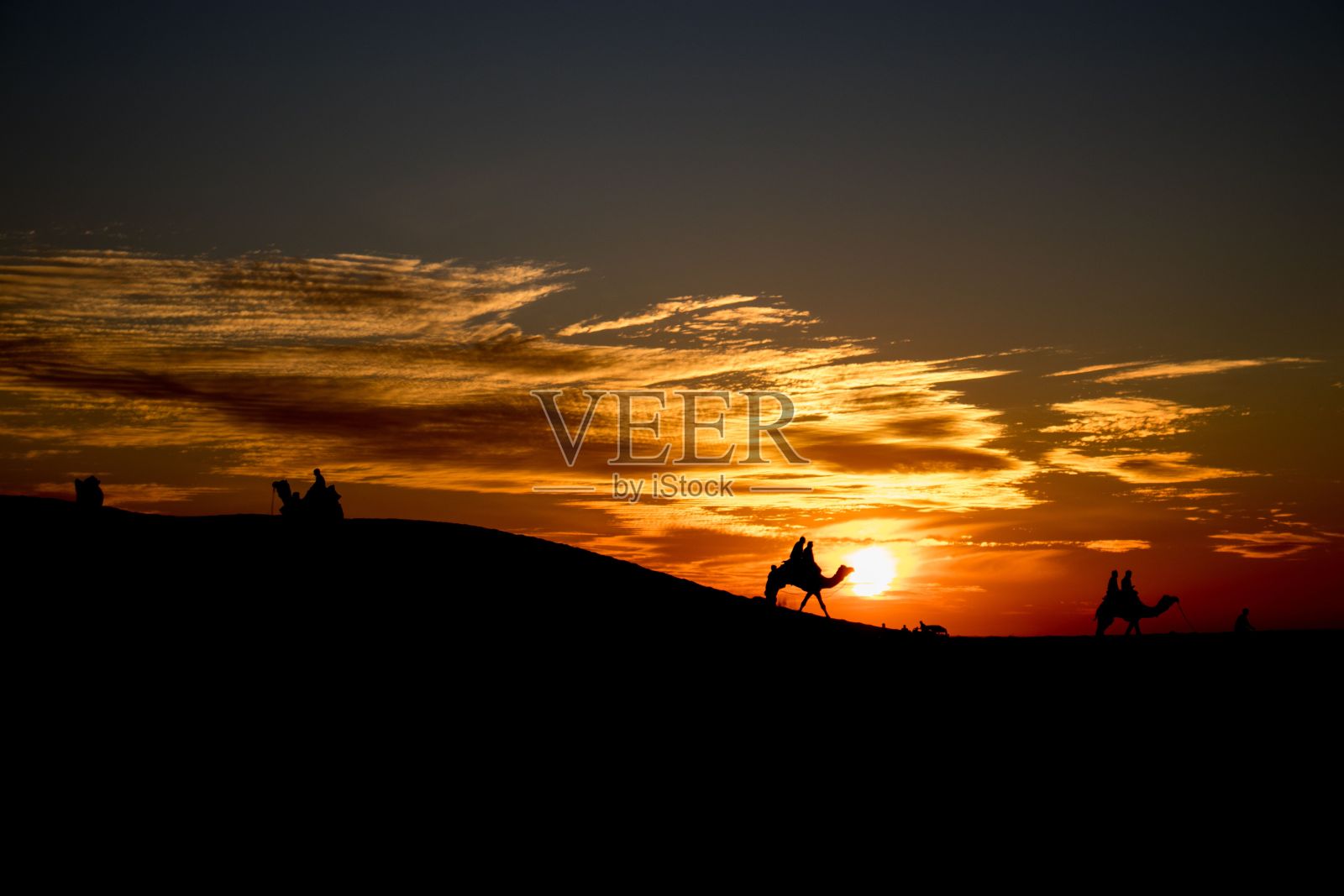 在橙色的夕阳下,人们和骆驼一起走过沙漠中的一座小山照片摄影图片