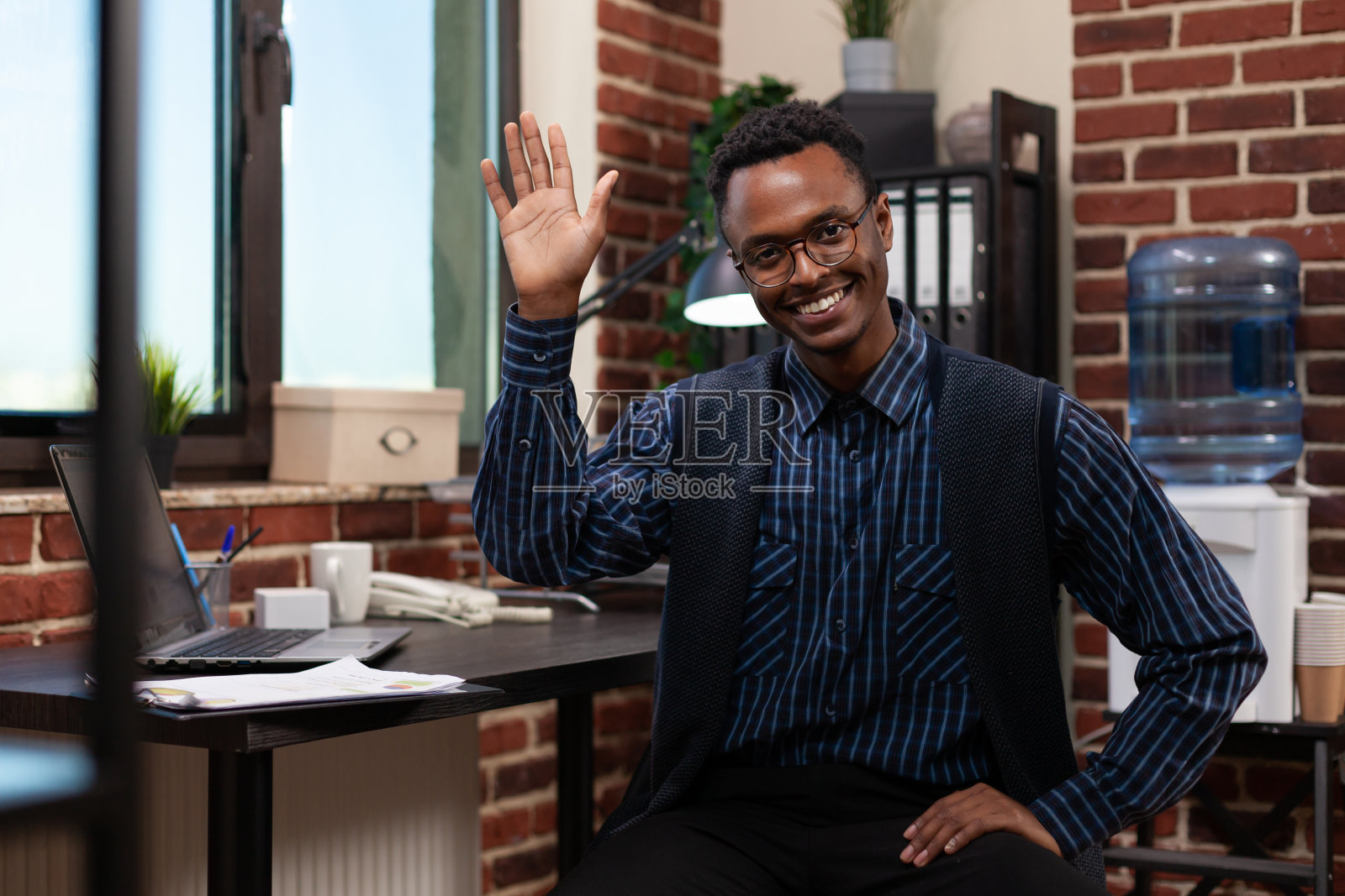 一名戴着眼镜、面带微笑、随意摆姿势、挥手打招呼的初创公司员工坐在办公桌前照片摄影图片