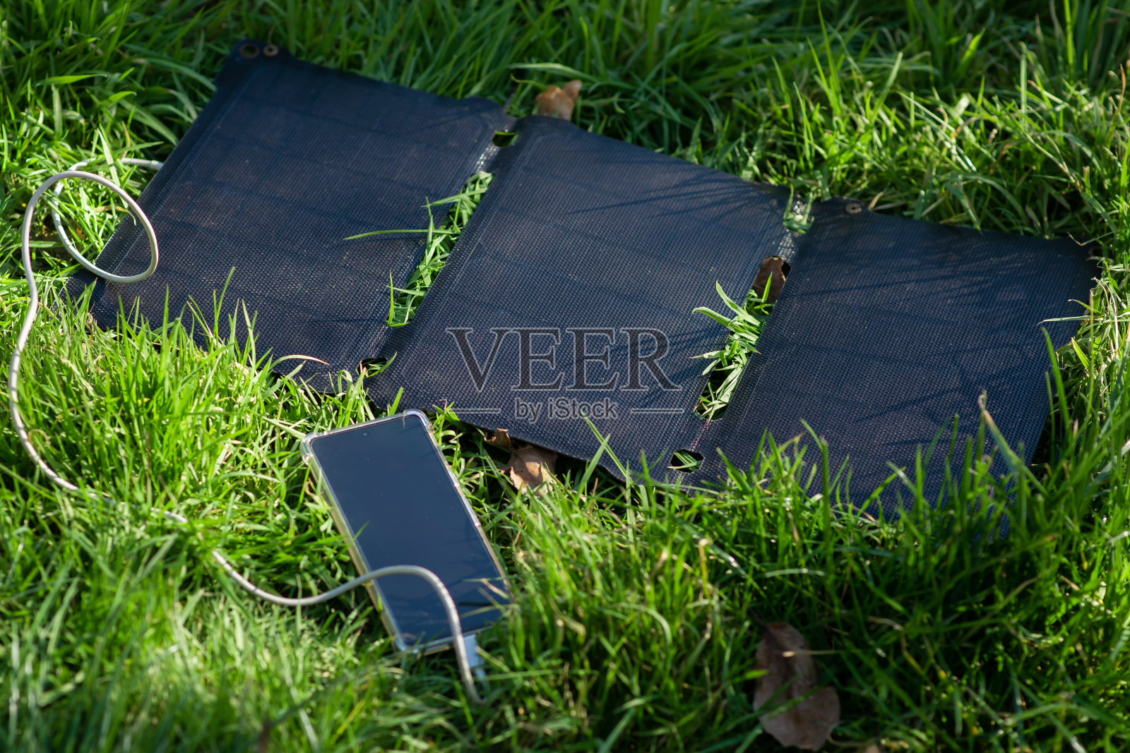 绿色草地上的便携式太阳能电池和手机。徒步旅行时,在户外娱乐时,给电子产品充电。生态能源概念。照片摄影图片