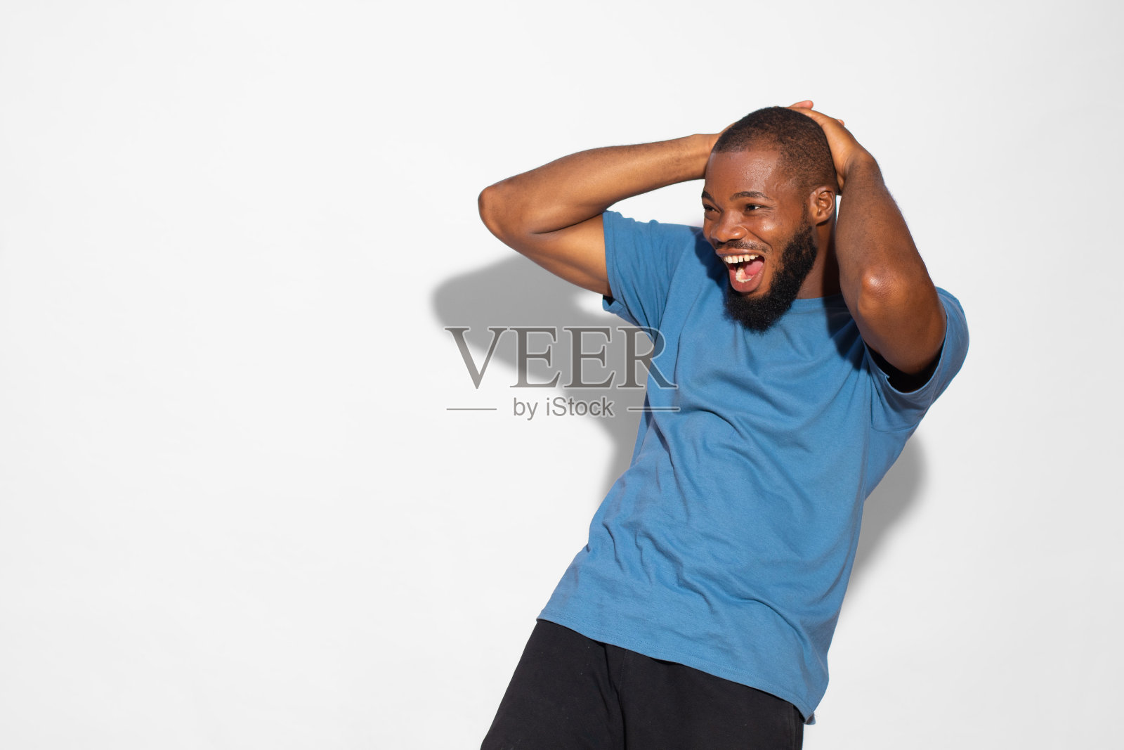 一个穿着蓝色t恤的黑人微笑着惊讶地双手放在白色背景的头上。照片摄影图片