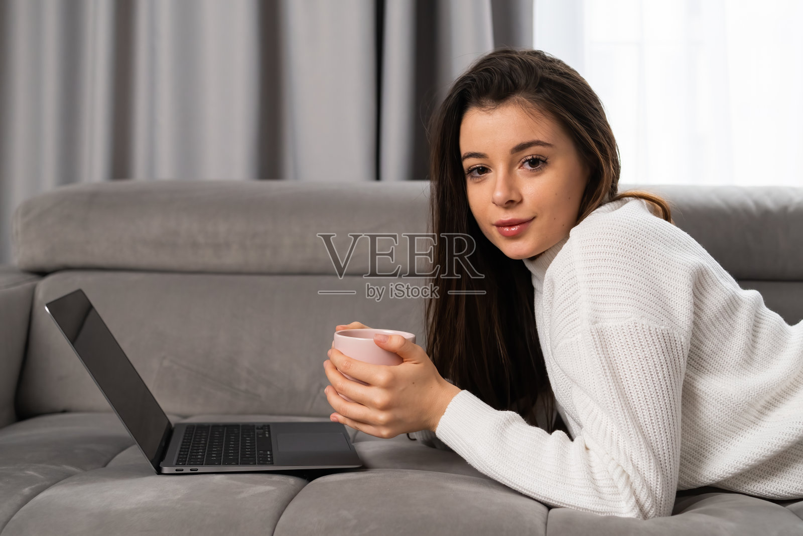 一个美丽的年轻女孩躺在沙发上,拿着一杯茶和一台笔记本电脑照片摄影图片