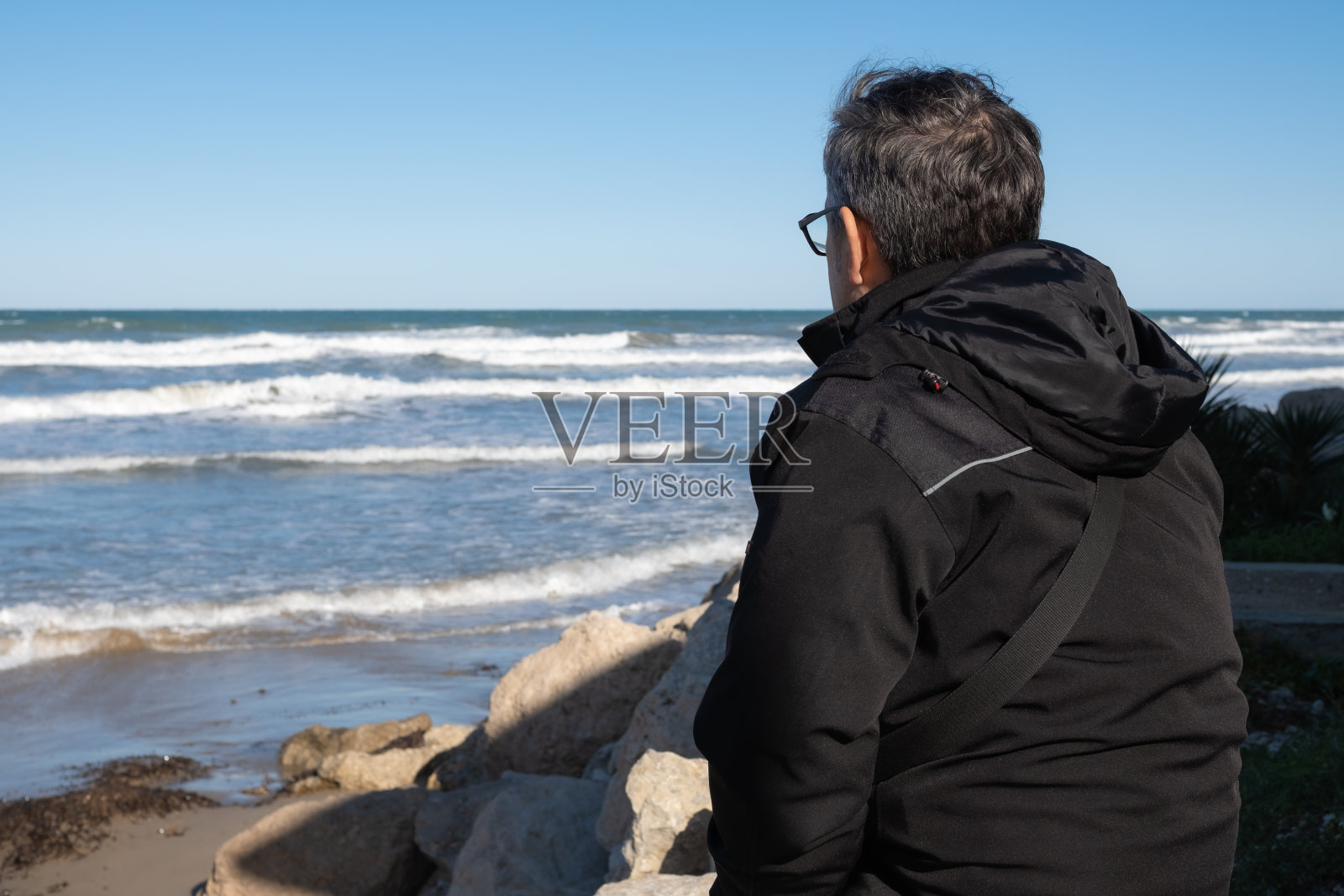 一个戴着眼镜、穿着深色外套的男人,在一个寒冷的冬日早晨,仰面坐在海滩的岩石上,若有所思地看着地中海。水平组合。Denia,阿利坎特省。照片摄影图片