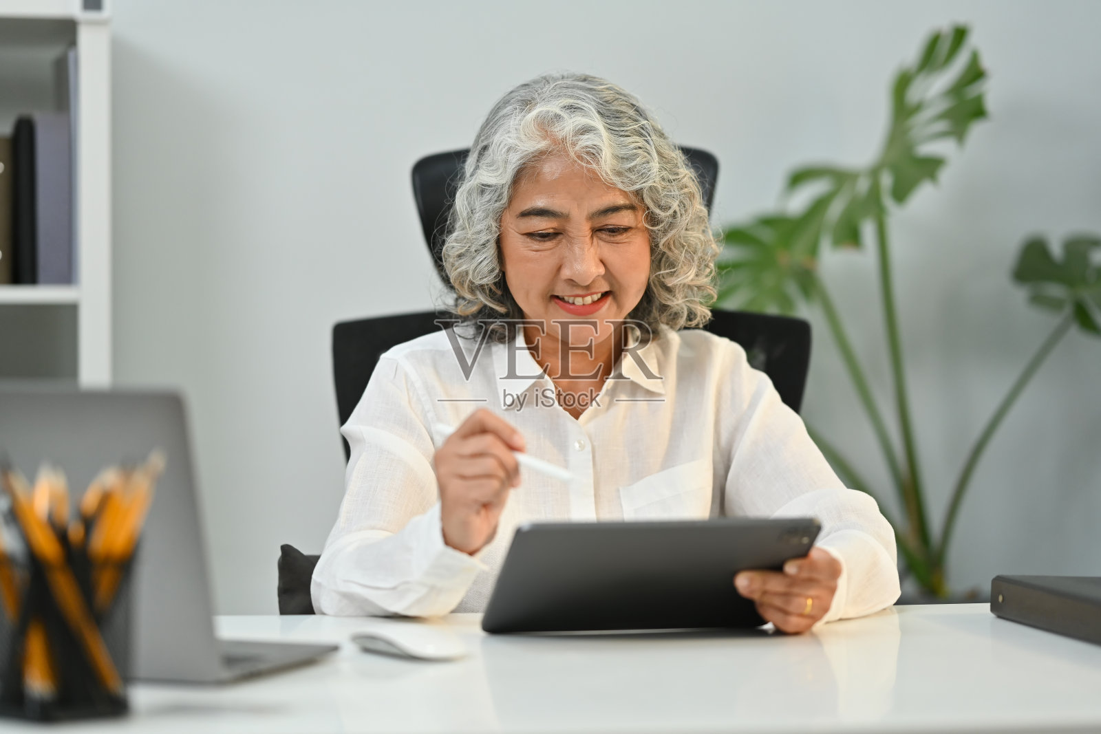 满意的中年妇女在家里办公室的平板电脑上浏览无线互联网。老年科技概念。照片摄影图片