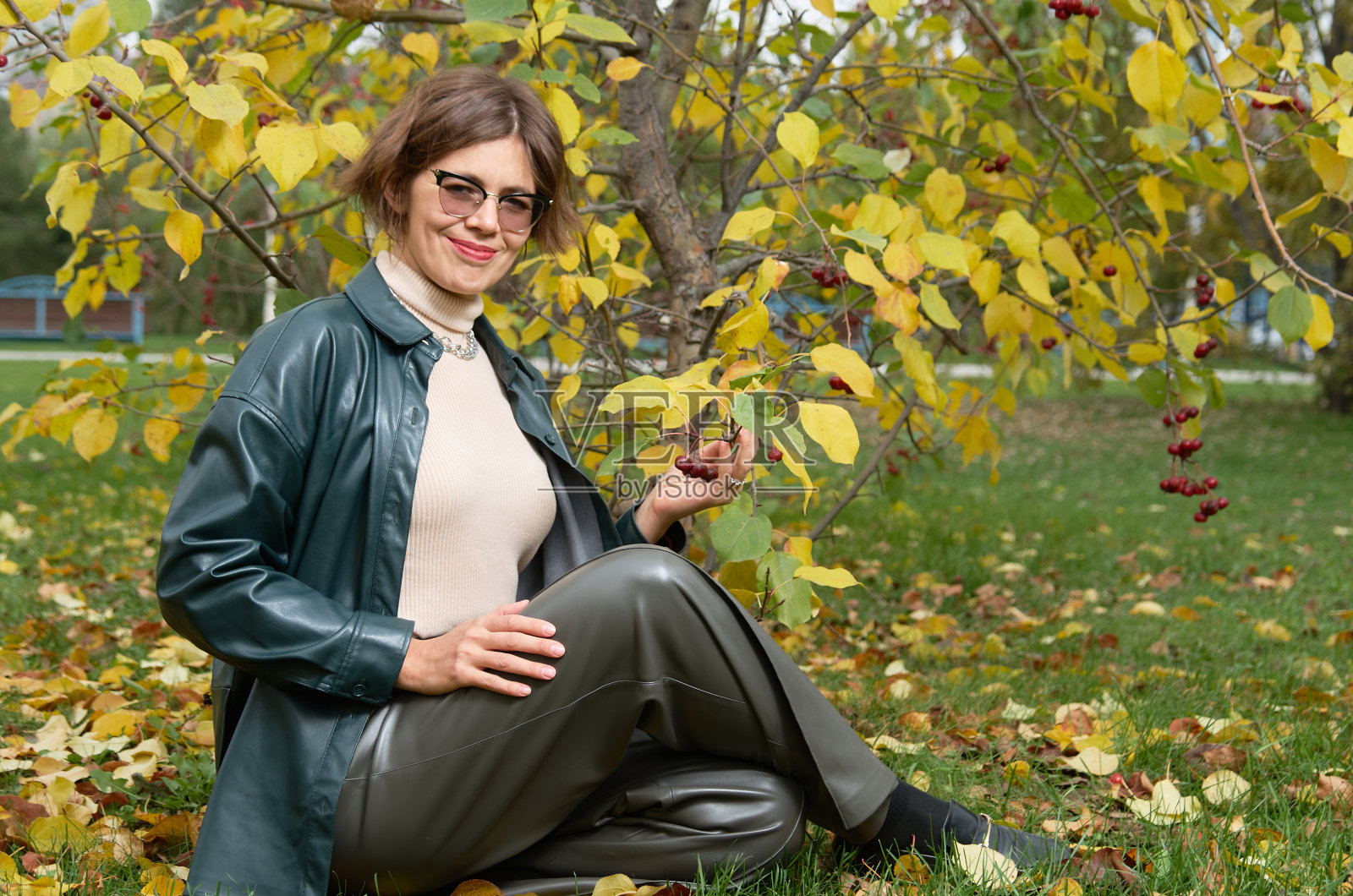 在秋天的公园里,一个穿着皮衣的女人坐在黄叶中间照片摄影图片