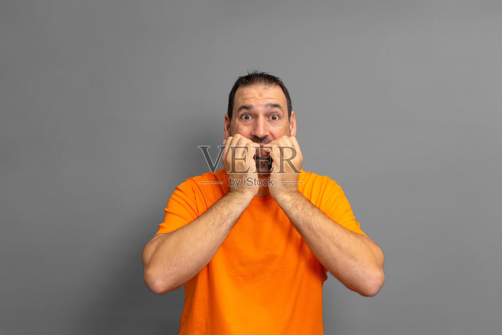 一名穿着橙色t恤、留着胡子的拉丁裔男子因为恐惧而咬着指甲,他正在经历一种痛苦的情况。孤立在灰色背景上。照片摄影图片