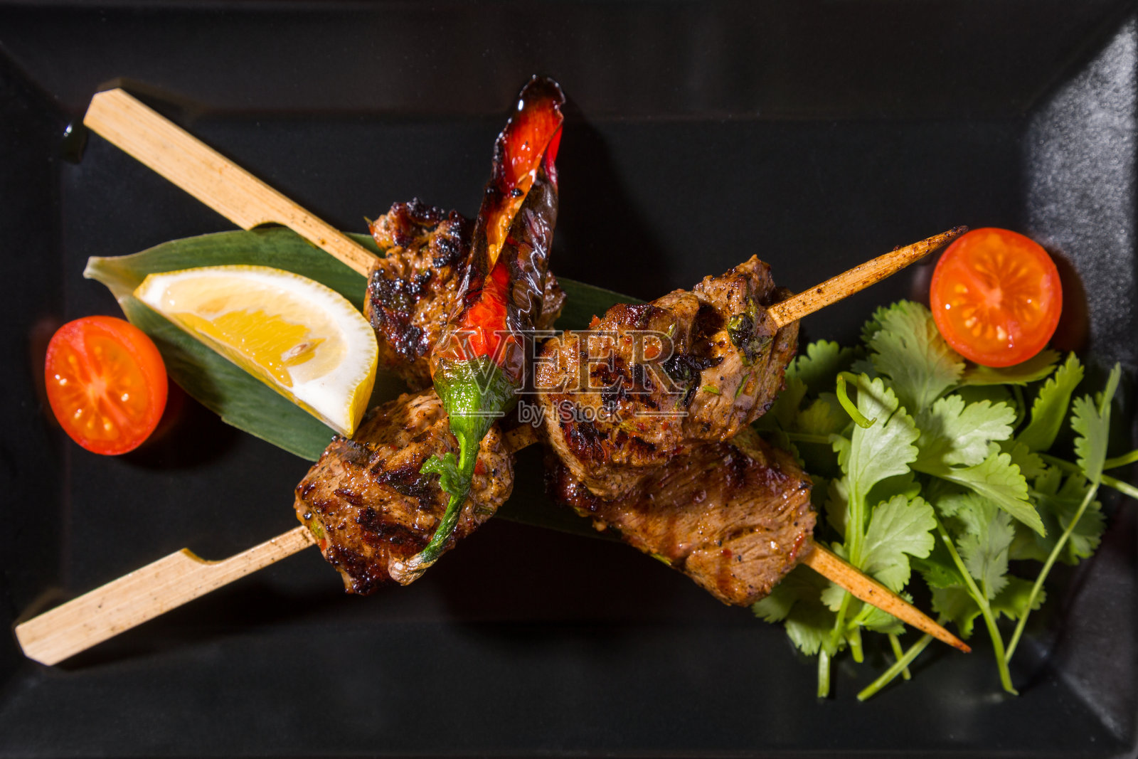 把烤肉串串在烤串上,用欧芹、番茄、柠檬和炸胡椒粉串在黑色的叶子上照片摄影图片