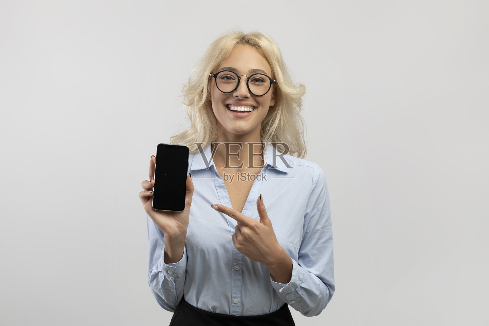 商务应用程序。快乐的商务女士手持智能手机,手指指向空白屏幕,浅灰色的工作室背景照片摄影图片
