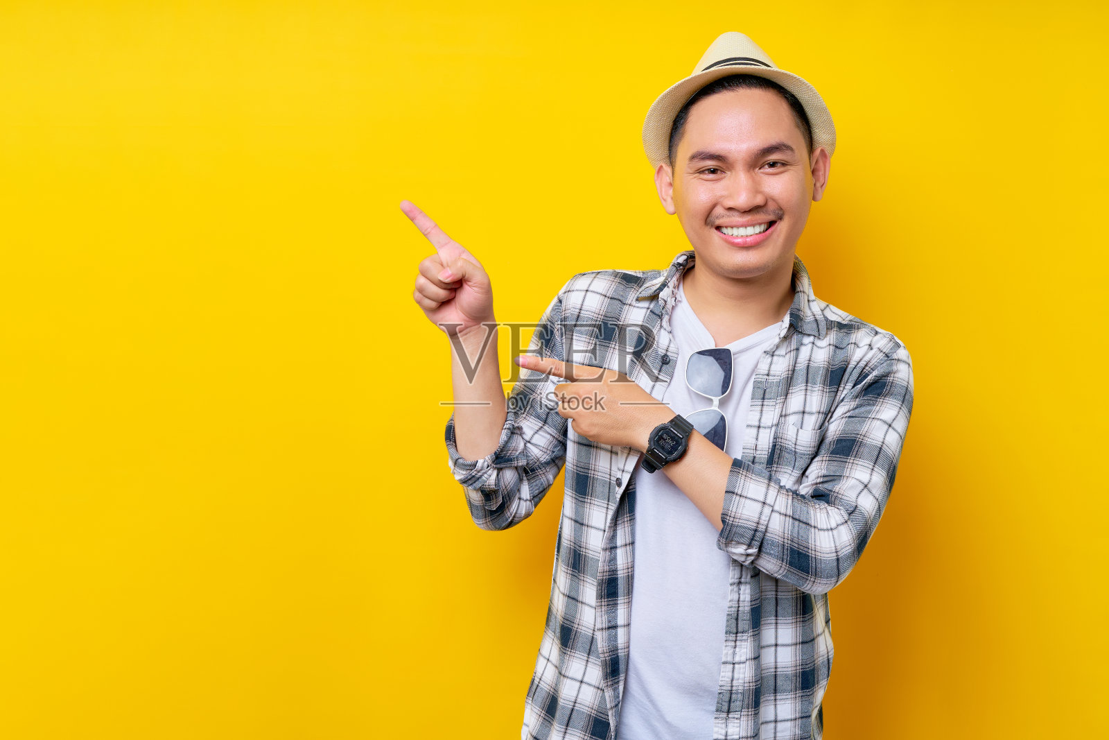 微笑开朗年轻英俊的亚裔男子20多岁,穿着休闲服,帽子站在一边指着手指在模拟复制空间隔离在黄色背景。人的生活方式概念照片摄影图片