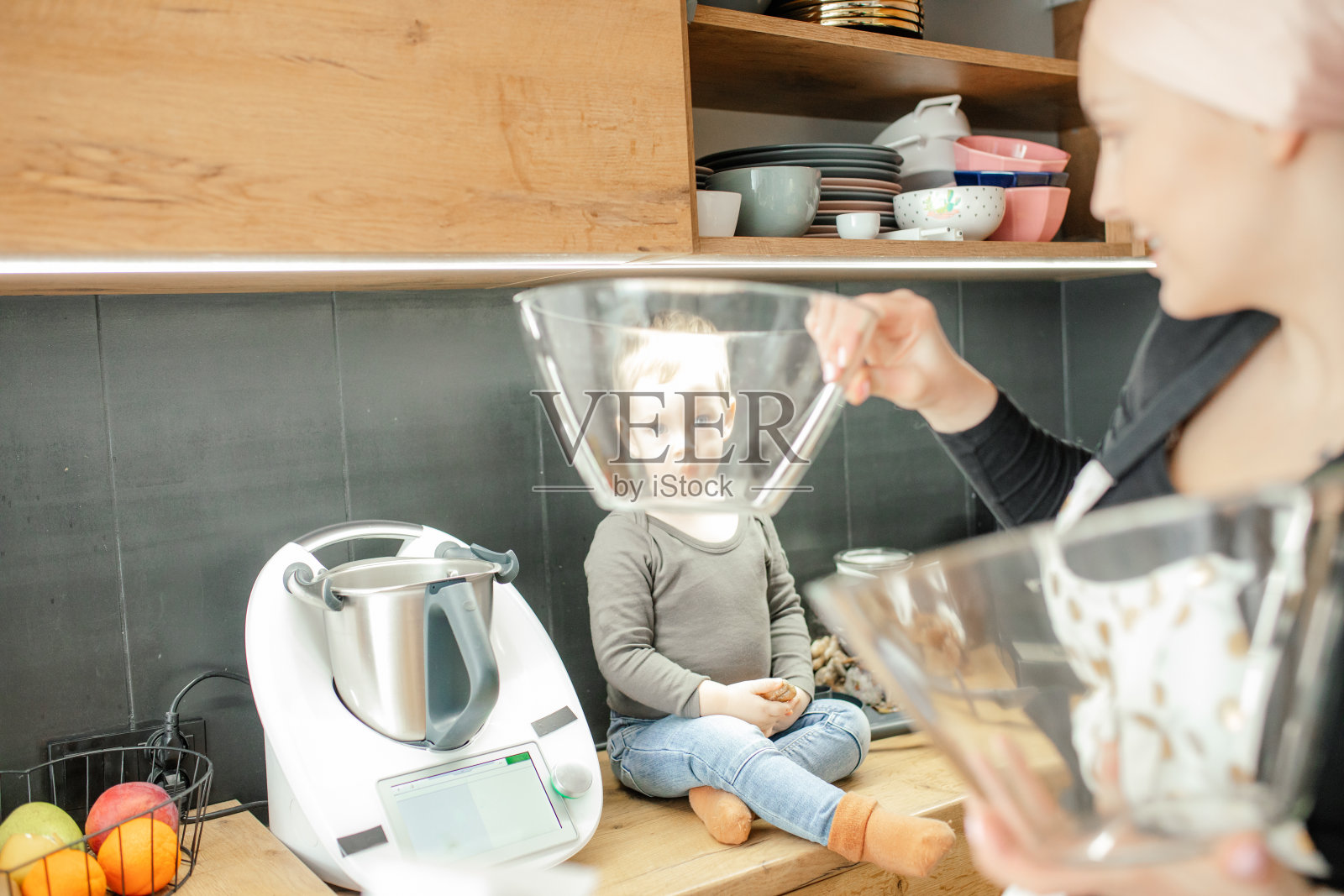 漫画,惊人的可爱的金发男孩坐在木制厨房抽屉,看着相机通过透明的碗在女人的手中照片摄影图片