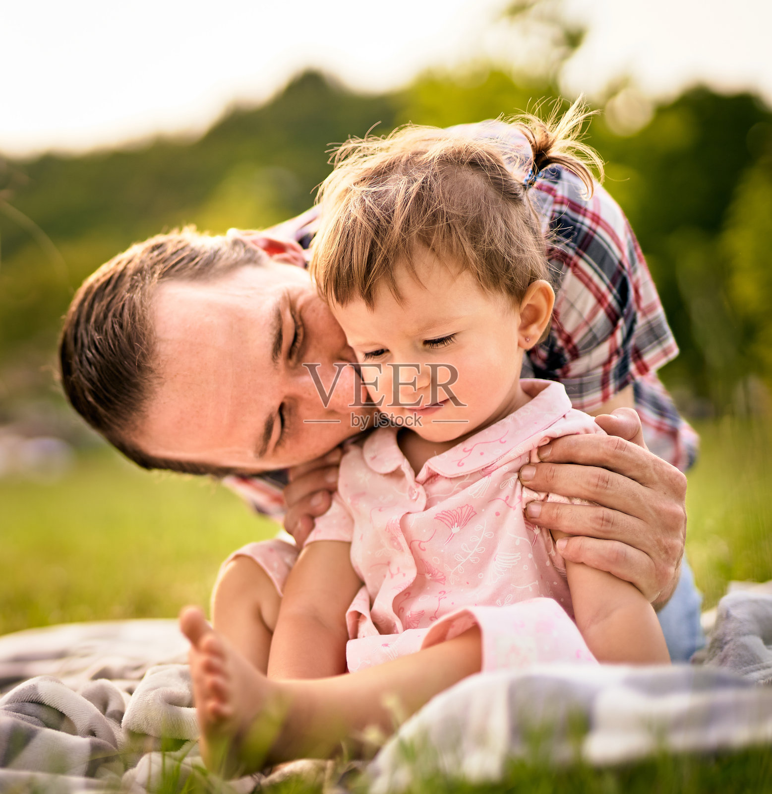 年轻的爸爸拥抱和亲吻他蹒跚学步的女儿- 20多岁的男人和他的孩子在户外玩耍-父母和童年的概念照片摄影图片