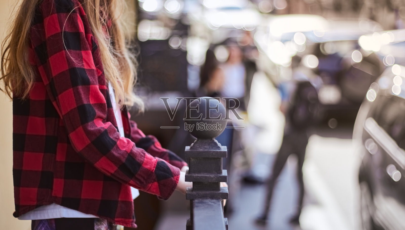 一个身穿红黑相间衬衫、金发长发的女孩正抓着栏杆。十几岁的女孩站在栏杆旁。女孩扶着扶手。照片摄影图片