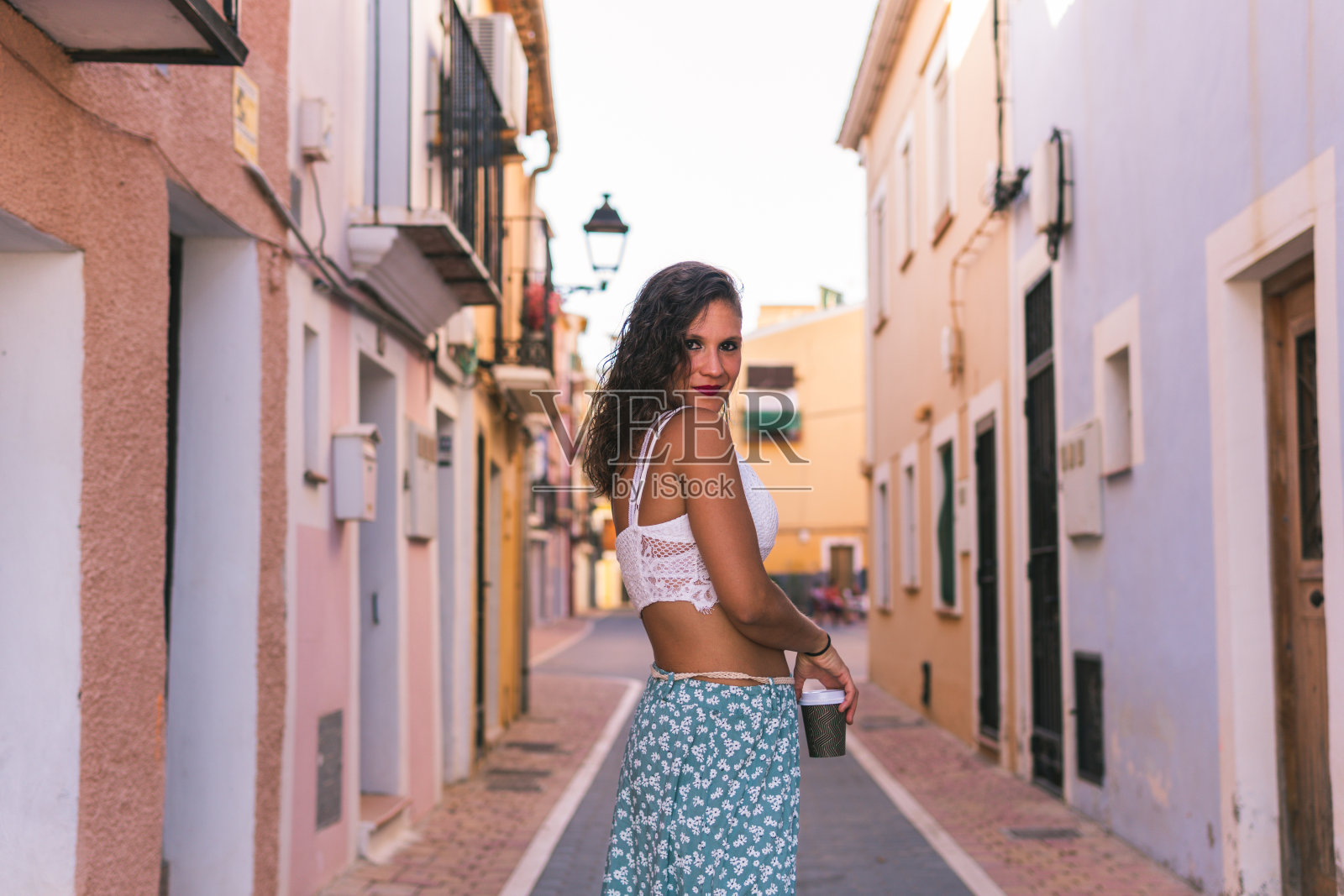 在西班牙阿利坎特的维拉约奥萨,五彩缤纷的街道上,一位微笑的妇女手持便携式咖啡杯,呼吸着新鲜空气照片摄影图片