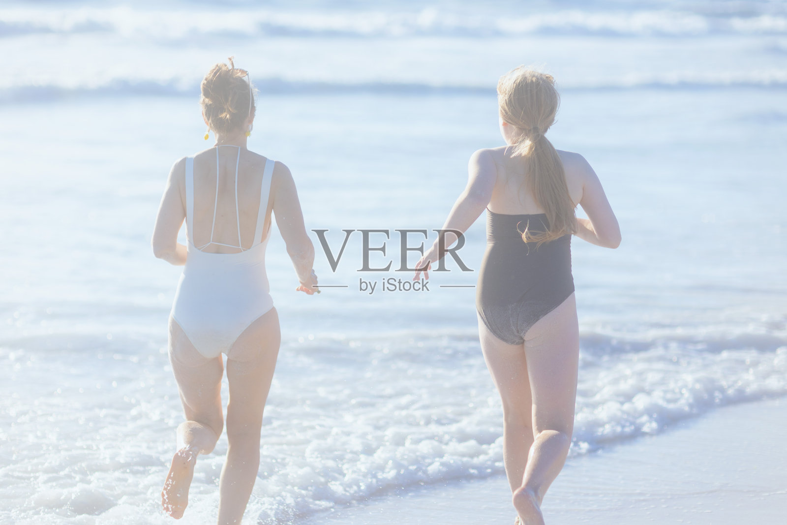 从后面看到母亲和十几岁的女儿在海滩上跑步照片摄影图片