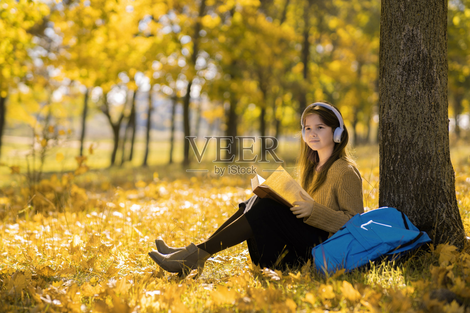 阳光明媚的秋日,快乐的女学生,戴着无线耳机,读着有趣的书,坐在公园的树叶上照片摄影图片