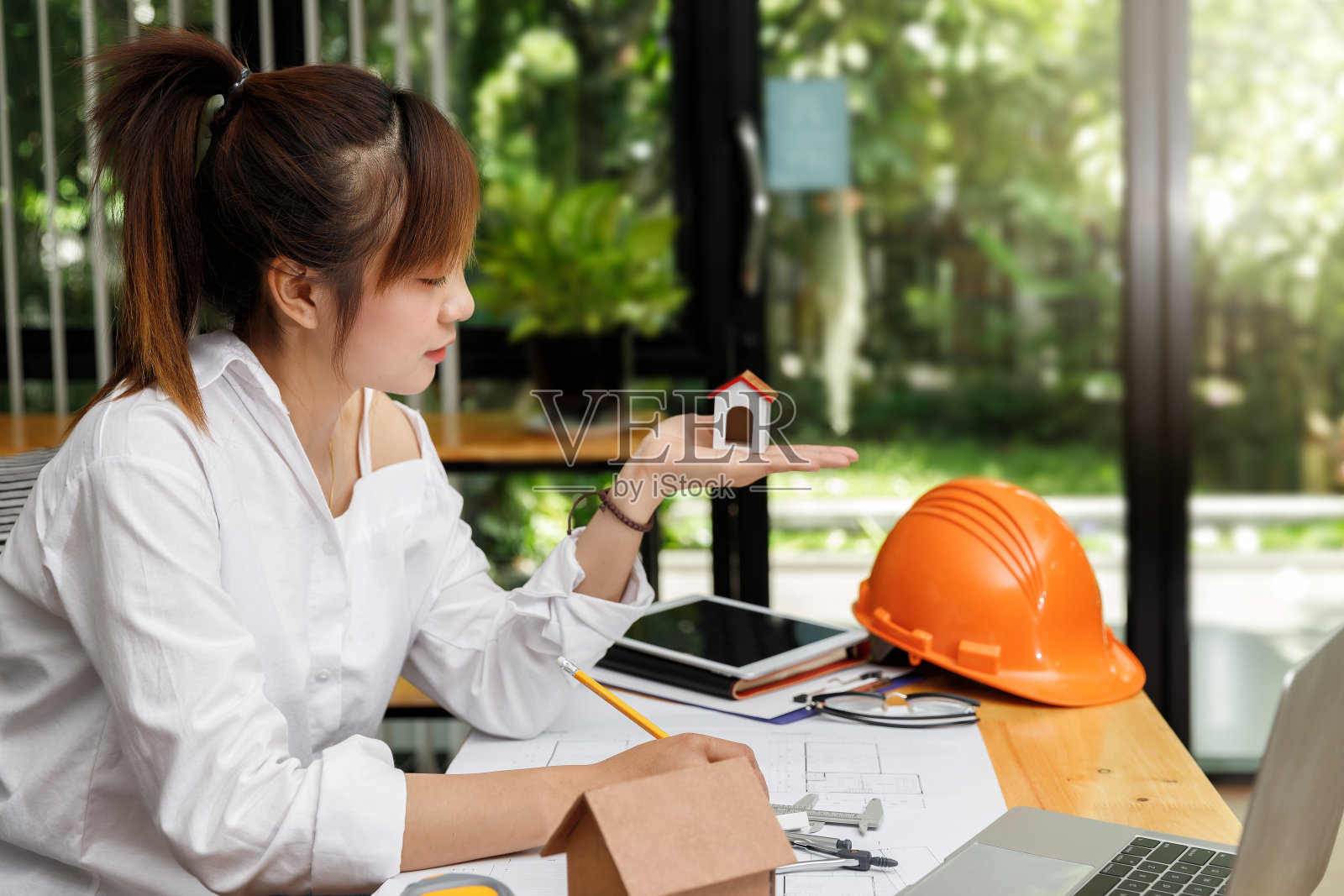 年轻漂亮的亚洲女人抱着房子模型,坐在她的工作地点前面。照片摄影图片