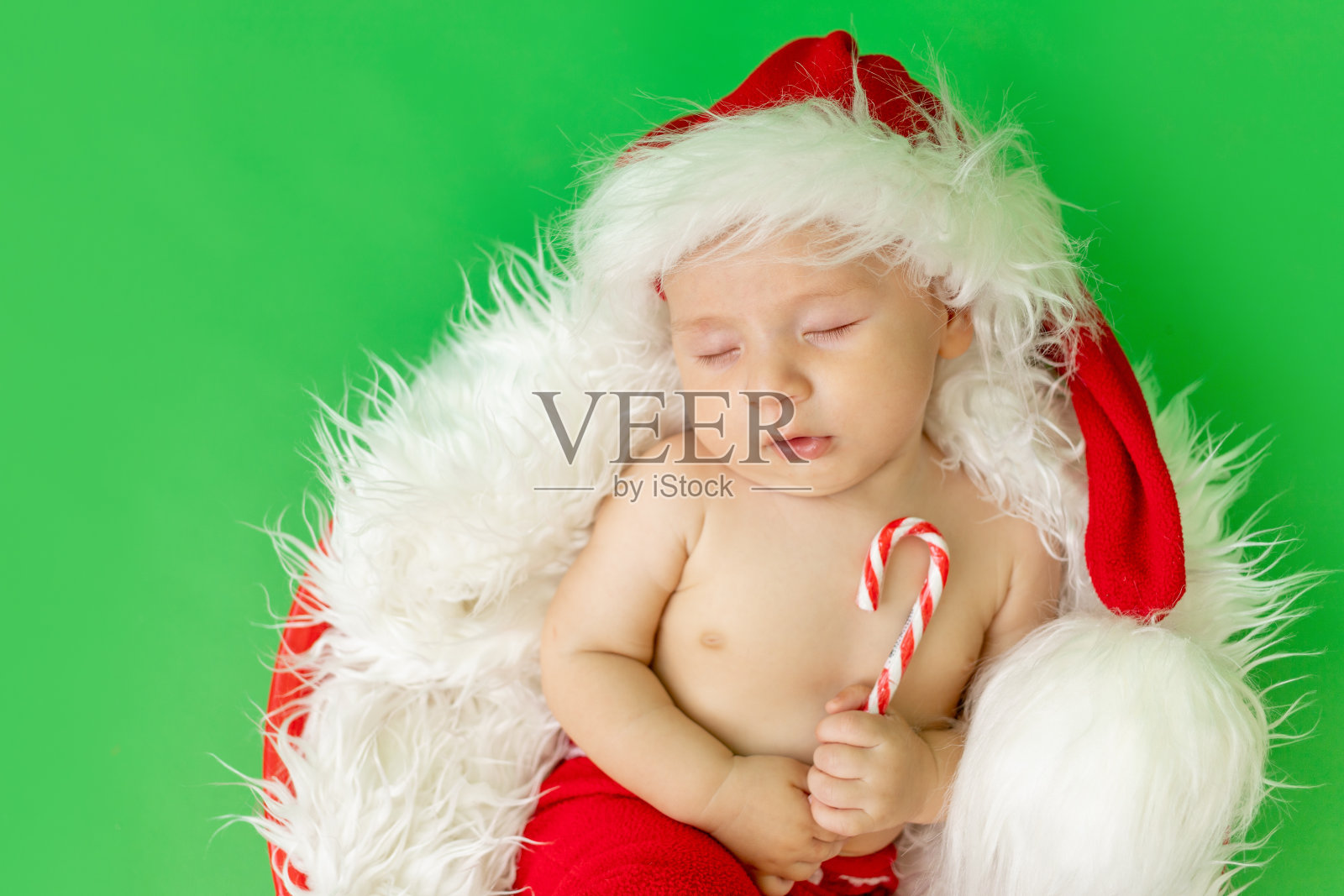一个穿着圣诞老人服装的熟睡的孩子,手里拿着一颗糖果,躺在绿色的孤立背景上,等待着新的一年和圣诞节,有文字的空间照片摄影图片