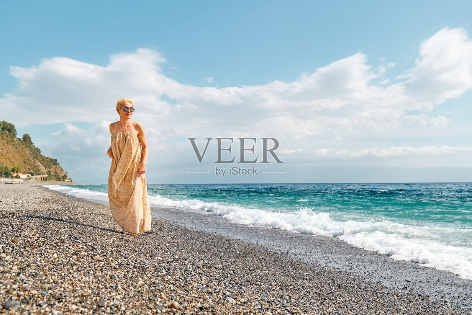 漂亮的金发女人走在卵石海滩上享受阳光多风的日子附近的蓝色大海。旅行的概念。柔和的颜色。照片摄影图片