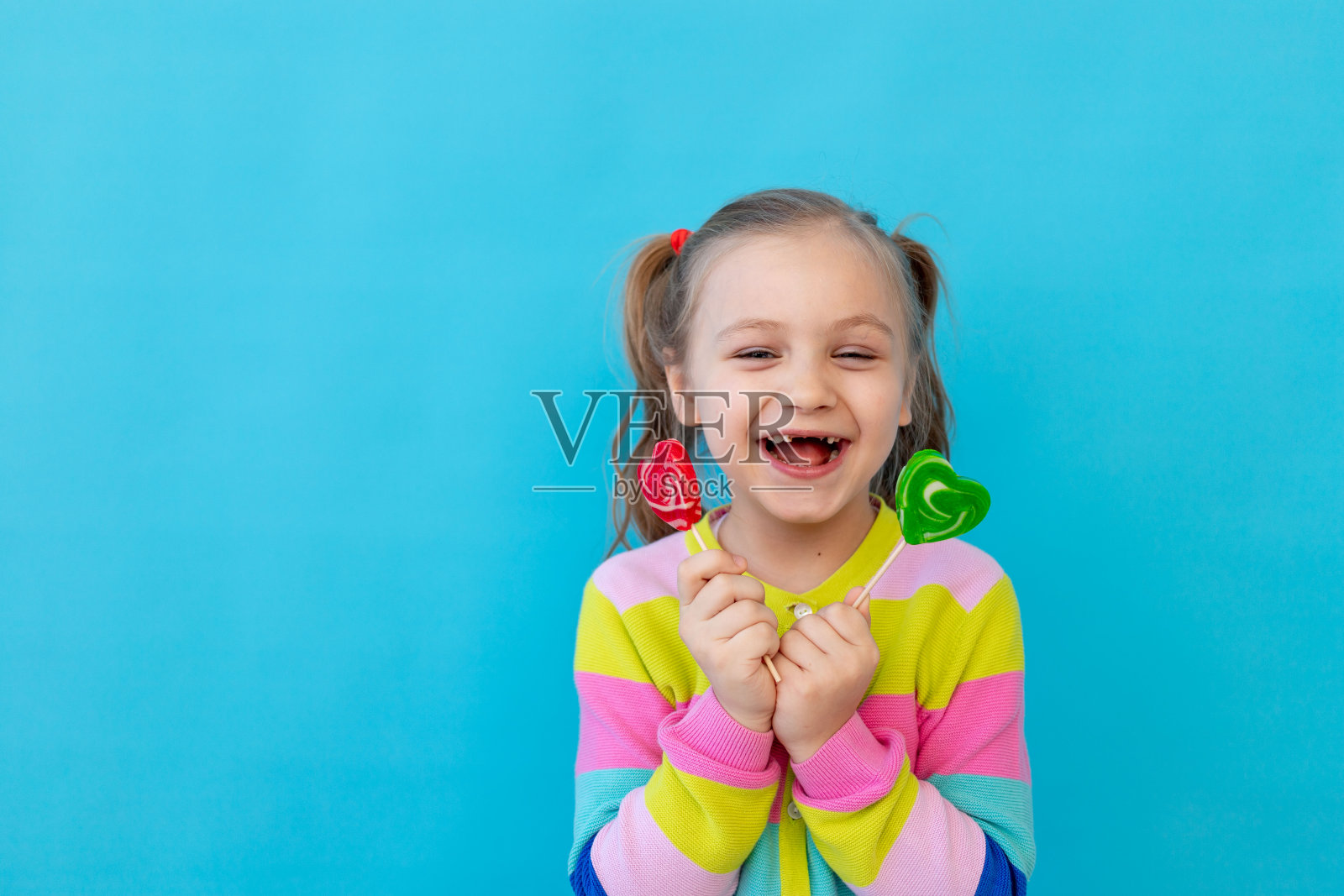 一个快乐的小女孩的肖像,在条纹夹克棒棒糖,一个大糖果棒。糖果和糖果的概念。蓝色背景,照相馆,文字的地方照片摄影图片