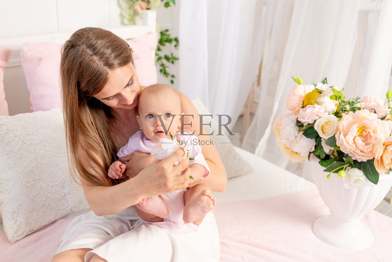 一位年轻美丽的母亲抱着6个月大的女儿坐在铺满鲜花的白色床上,拥抱着女儿,这是一个发短信的地方照片摄影图片