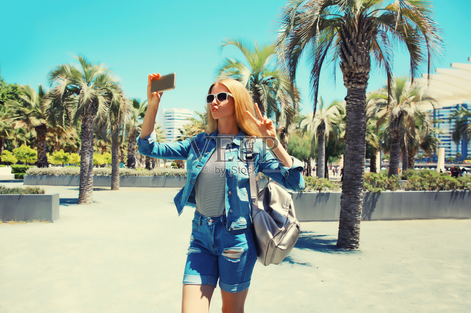 年轻女子在夏天公园用手机自拍的肖像,背着背包,背景是棕榈树照片摄影图片