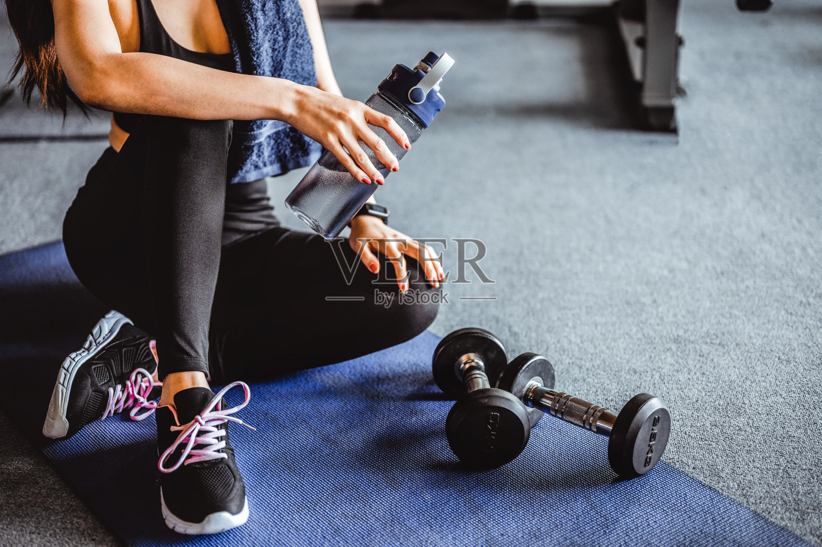 健身亚洲女子运动后放松,喝水和哑铃放在旁边的健身房。训练后放松。美丽的年轻女子看向别处,而坐在健身房。照片摄影图片