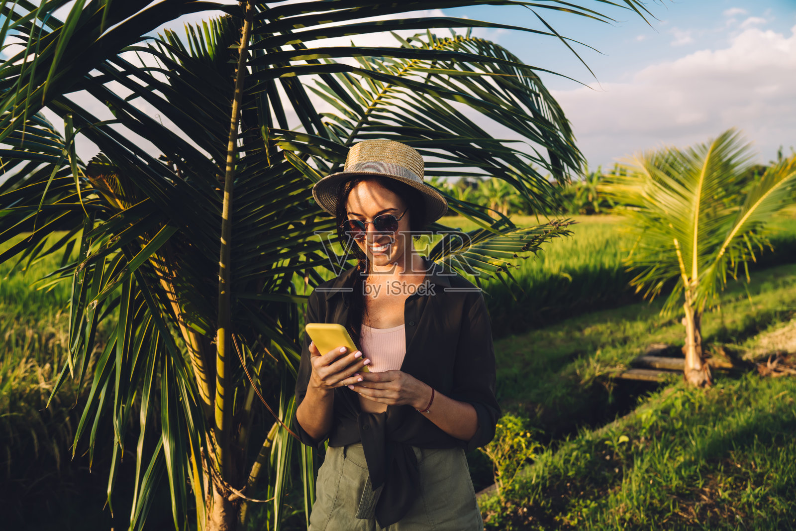 快乐的女游客在热带度假时享受智能手机短信,连接4g漫游发送短信,快乐的女人戴着太阳镜和草帽阅读移动出版物照片摄影图片