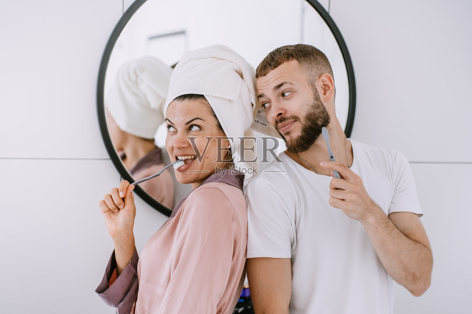 俏皮的西班牙年轻夫妇在浴室刷牙牙齿微笑拥抱。新婚夫妇在酒店房间享受蜜月。漂亮的白人女孩头上有毛巾和男朋友玩。照片摄影图片