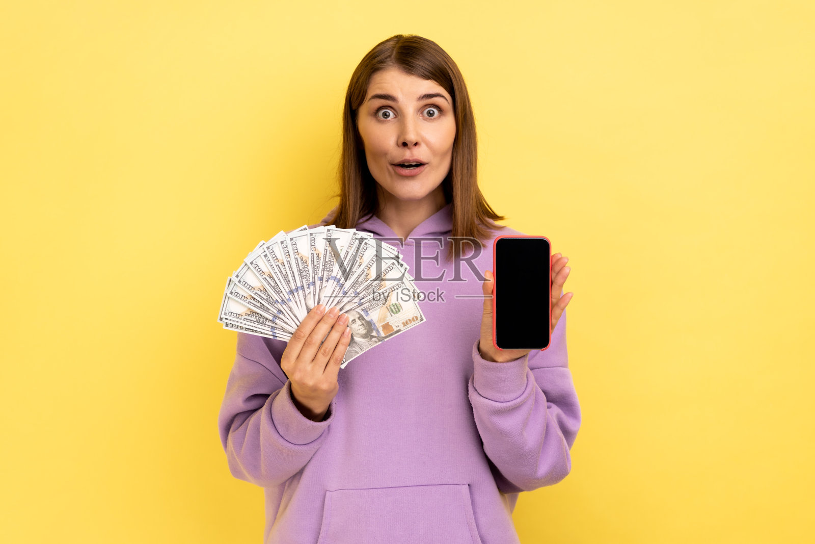 震惊的黑发女子拿着美元钞票和空屏幕的手机,在线赌博。照片摄影图片