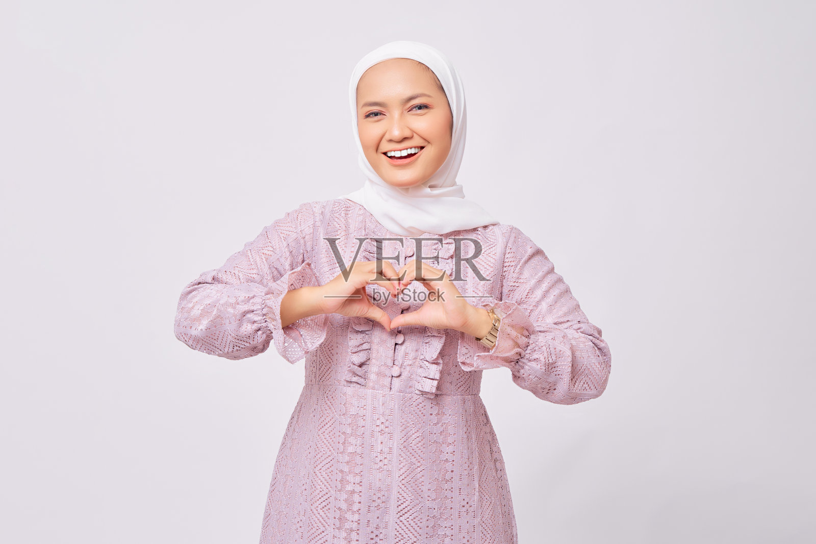 微笑的美丽年轻的亚洲穆斯林妇女戴头巾和紫色连衣裙显示心脏手势的肖像,使爱的标志与手指隔离在白色的工作室背景照片摄影图片