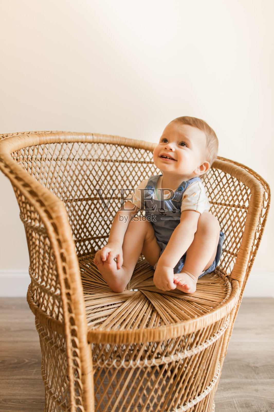 一个15个月大的古巴裔美国男婴,浅棕色短发,棕色眼睛和12只脚趾,穿着胡萝卜连体衣和蓝色兔子连体裤,还有蓬松的兔子尾巴,光着脚坐在复古藤椅上抬头看照片摄影图片